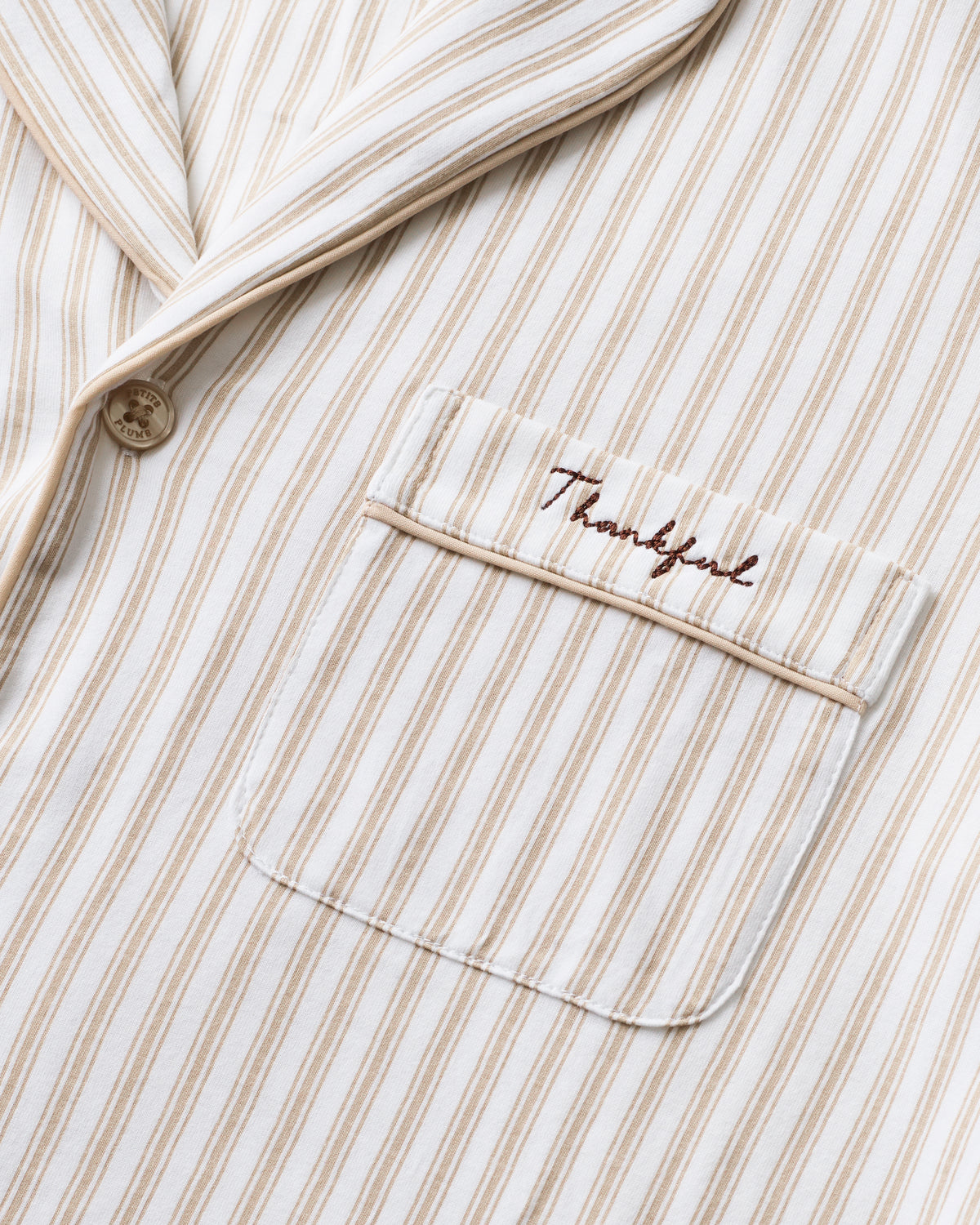 The Women's Pima Pajama Set in Camel Ticking with "Thankful" embroidery features a beige and white striped, long-sleeve button-up top with a chest pocket and matching elastic-waist pants, shown neatly arranged on a white background.