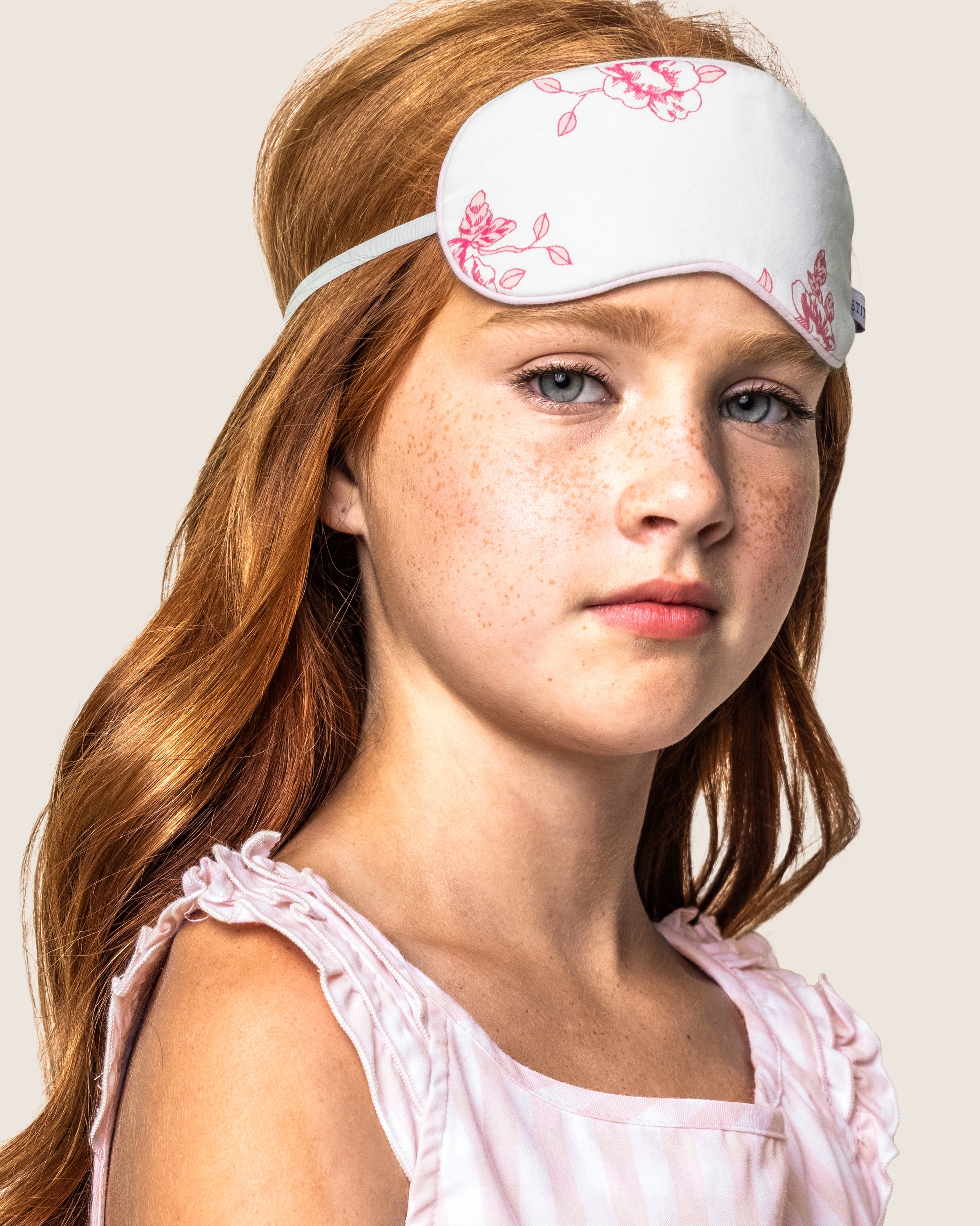 A young girl with long red hair gazes forward, wearing a Kids Sleep Mask in English Rose Floral by Petite Plume, with a pink sleeveless top against a neutral background. Perhaps this is the start of a memorable moment, just before gifting her matching eye masks to friends.