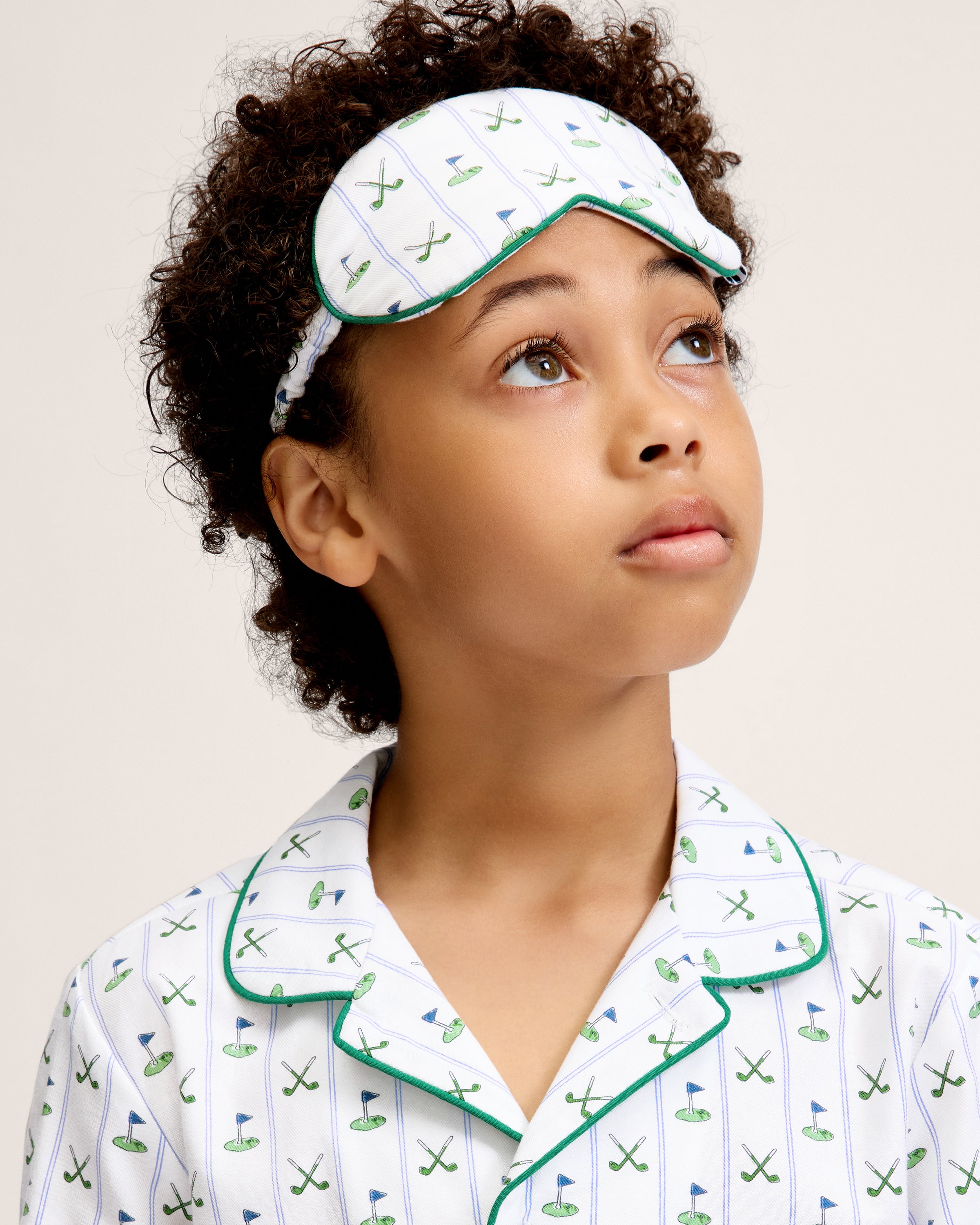 The Kid's Twill Sleep Mask in Fairway Stripe features a white design with green trim, decorated with golf clubs and flags. It includes an adjustable strap and a PETITE tag.