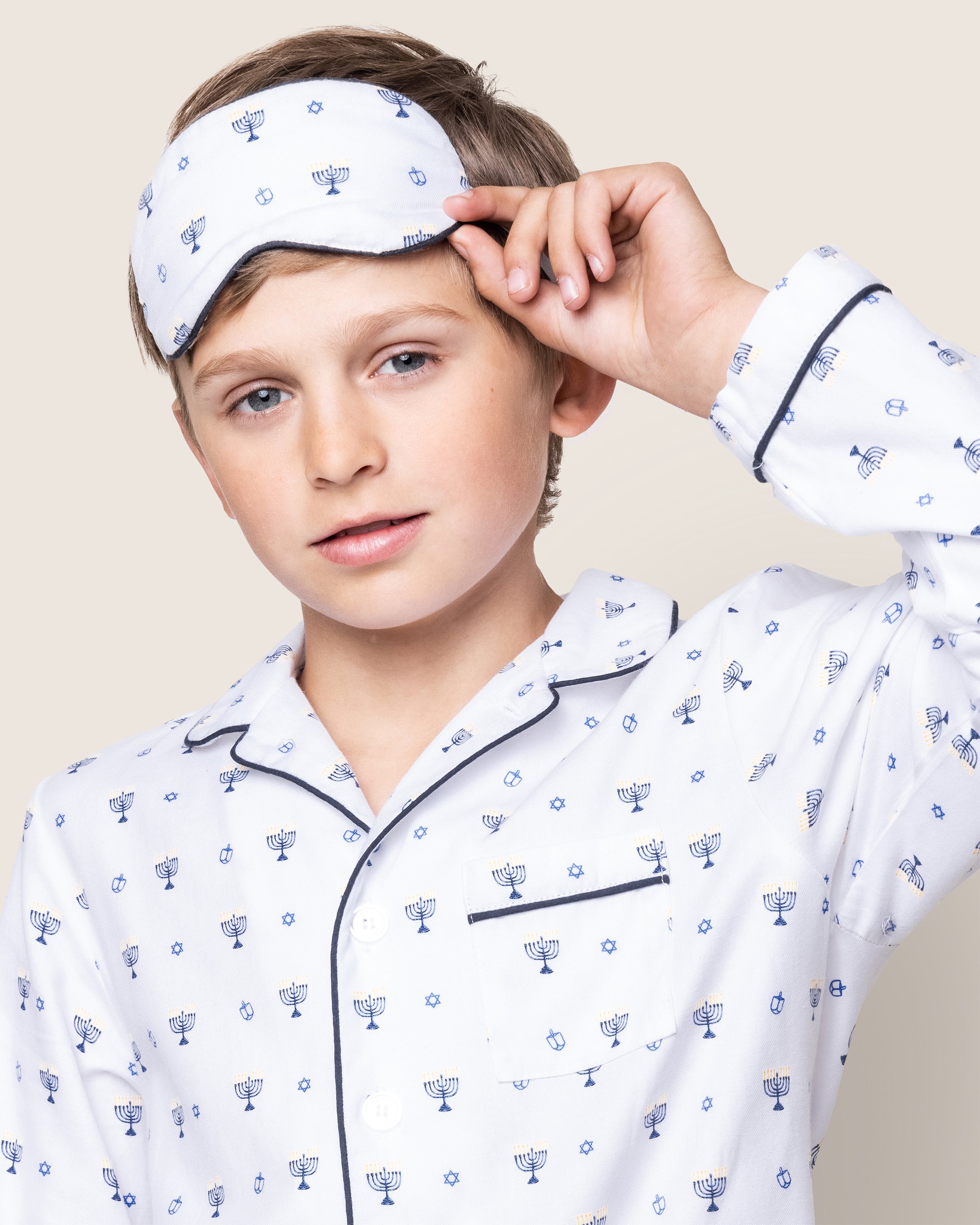 A child wears Hanukkah pajamas featuring a white set with blue menorah patterns and navy piping on the collar and pocket. They hold Petite Plumes Kids Twill Sleep Mask in Happy Hanukkah above their eyes, gazing forward against a plain background.