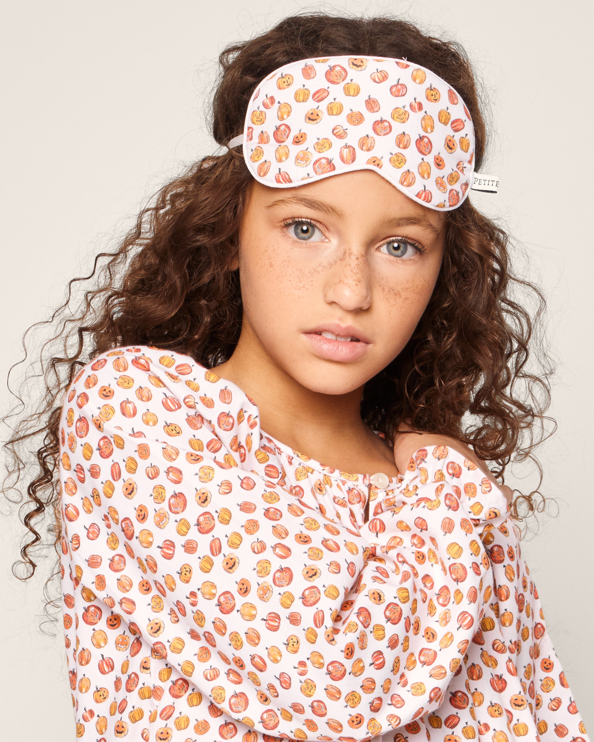 A girl with curly brown hair models matching Petite Plume pajamas and the Kids Twill Sleep Mask in Pumpkin Patch against a neutral background, her festive attire celebrating a Halloween print as she gazes toward the camera.