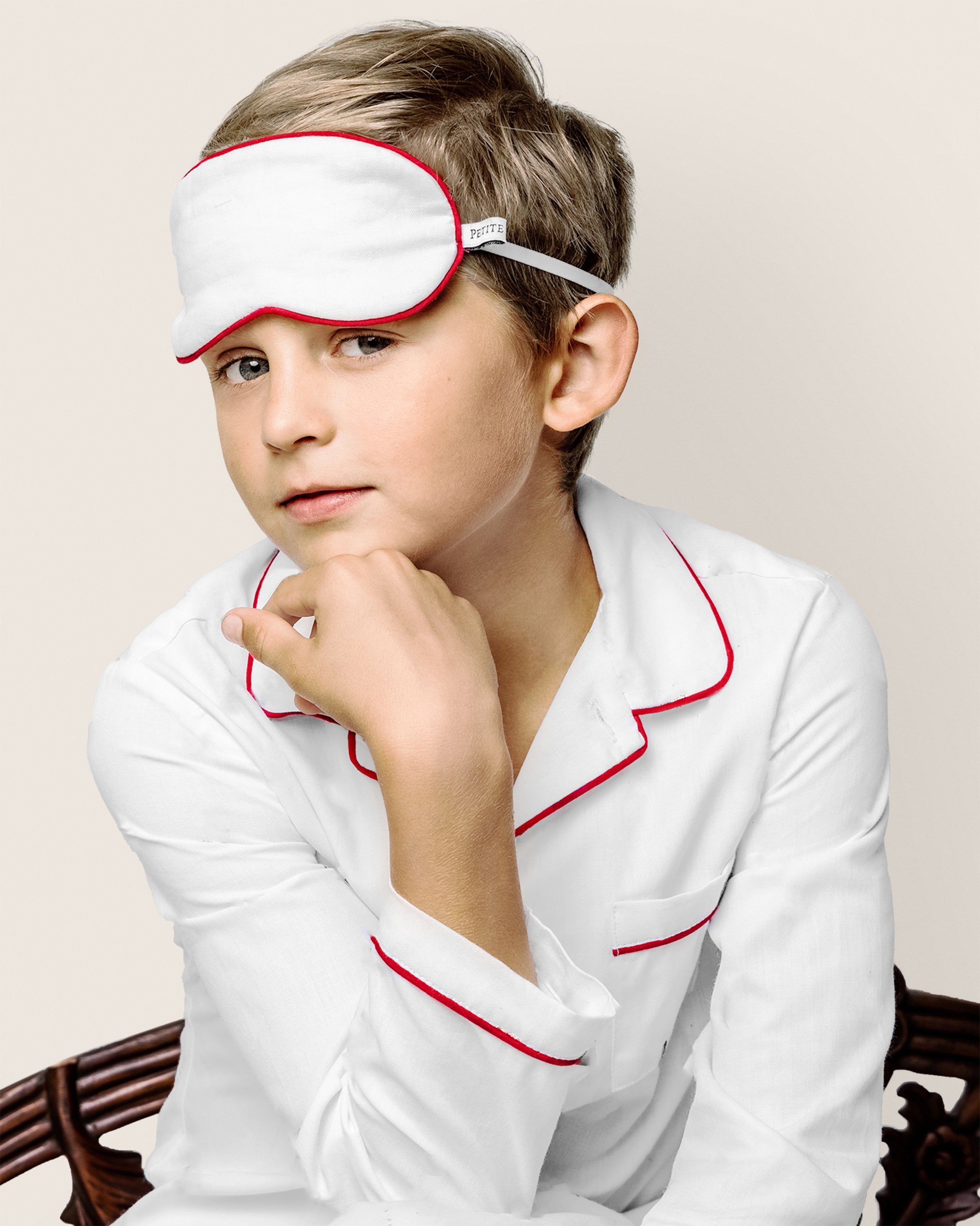 A child in Petite Plumes luxurious Kids Twill Pajama Set and matching White with Red Piping Sleep Mask rests their chin on a wooden chair against a plain background.