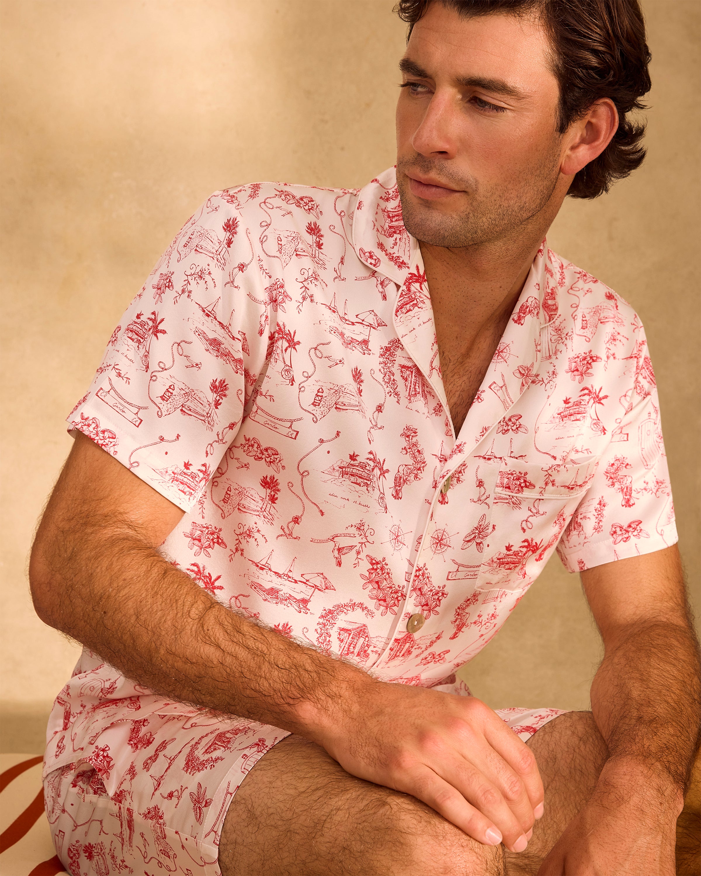 The Petite Plume x Eden Rock Reverie Men's Silk Short Set includes a short-sleeve button-up top and matching shorts with an elastic waistband, featuring a white base and red toile-style print with intricate scenes.