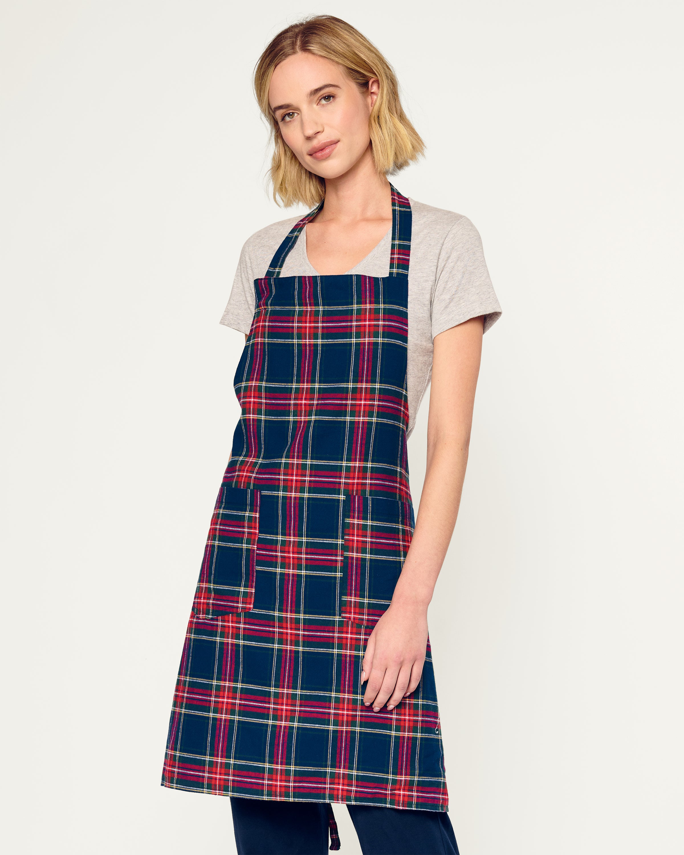 Two Adult Aprons in Windsor Tartan by Petite Plume, ideal for holiday baking, hang on a wooden rack beside a striped towel. One apron is labeled Dad, with a wooden spoon in its pocket. The background features a cozy warm beige wall.