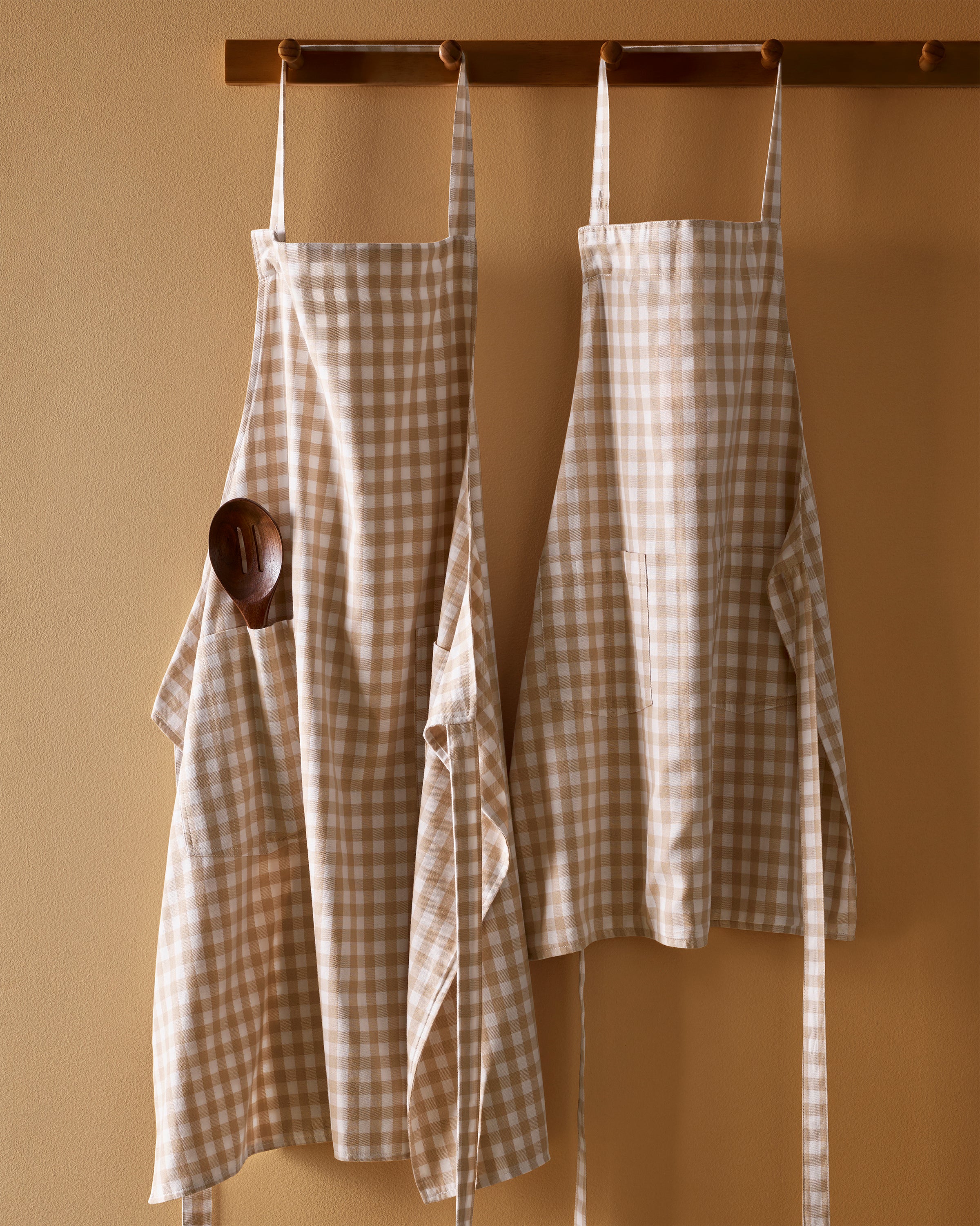 The Kid's Twill Apron in Camel Gingham, featuring a beige and white gingham pattern, two front pockets, and long waist ties, is displayed on a white background.