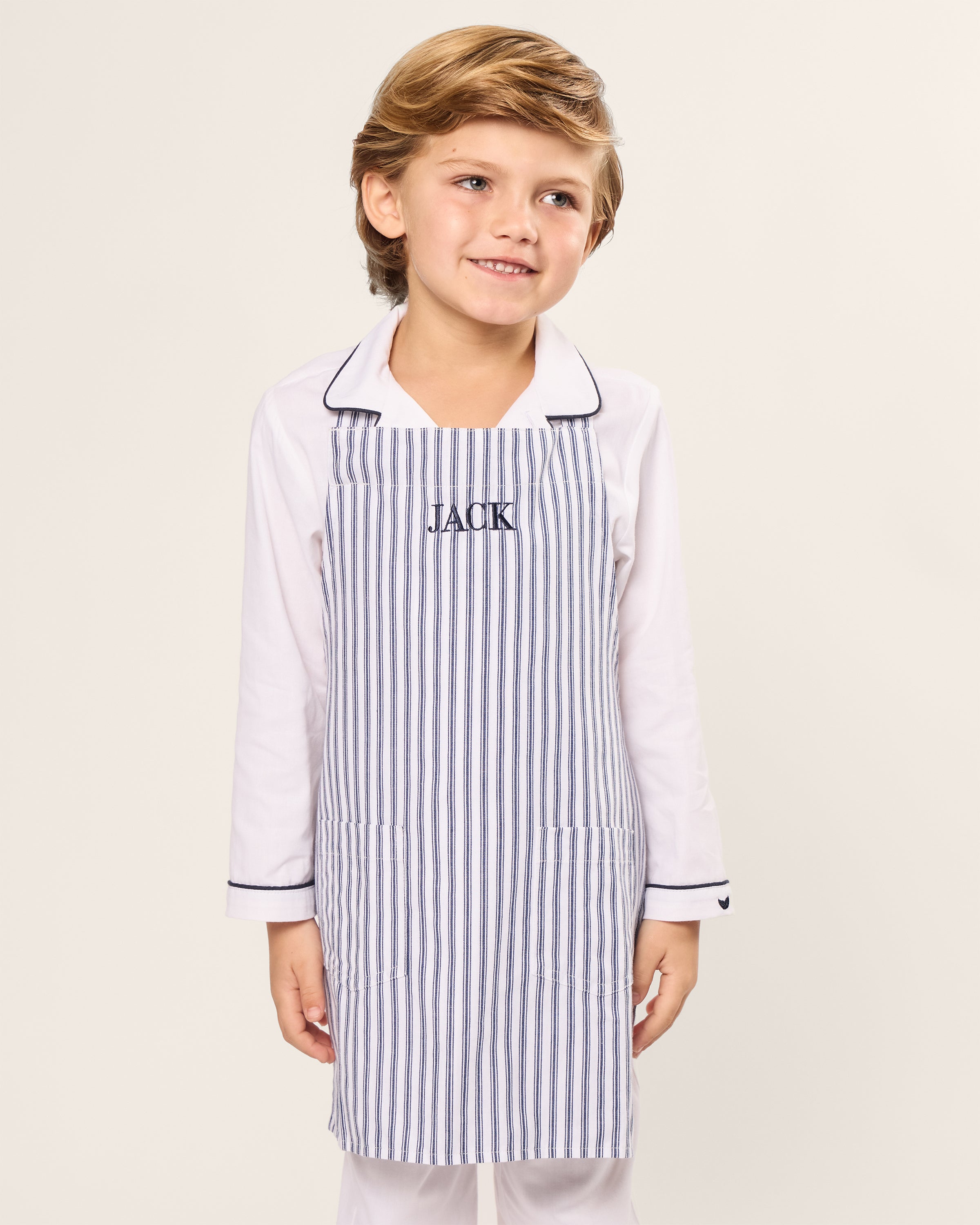 A young child with light brown hair wears a childrens size white outfit and the Kids Apron in Navy French Ticking, embroidered with JACK, from Petite Plume. They beam cheerfully against a plain background, ready for some holiday baking fun!.