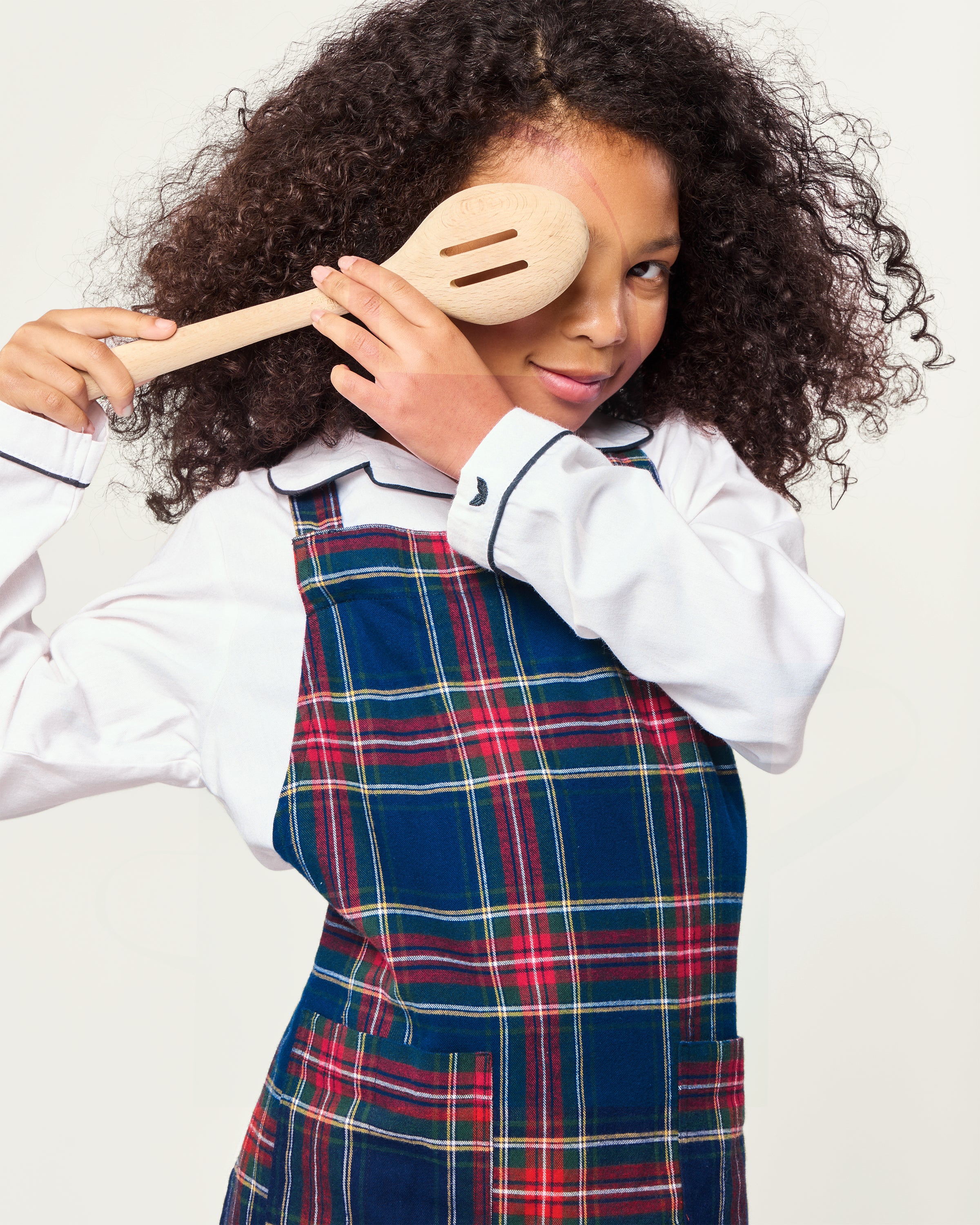 Two Windsor tartan kids aprons from Petite Plume hang on a wooden rack next to a striped dish towel, evoking holiday baking nostalgia. One apron, embroidered with Dad, holds a wooden spoon in its pocket against the warm beige wall backdrop.