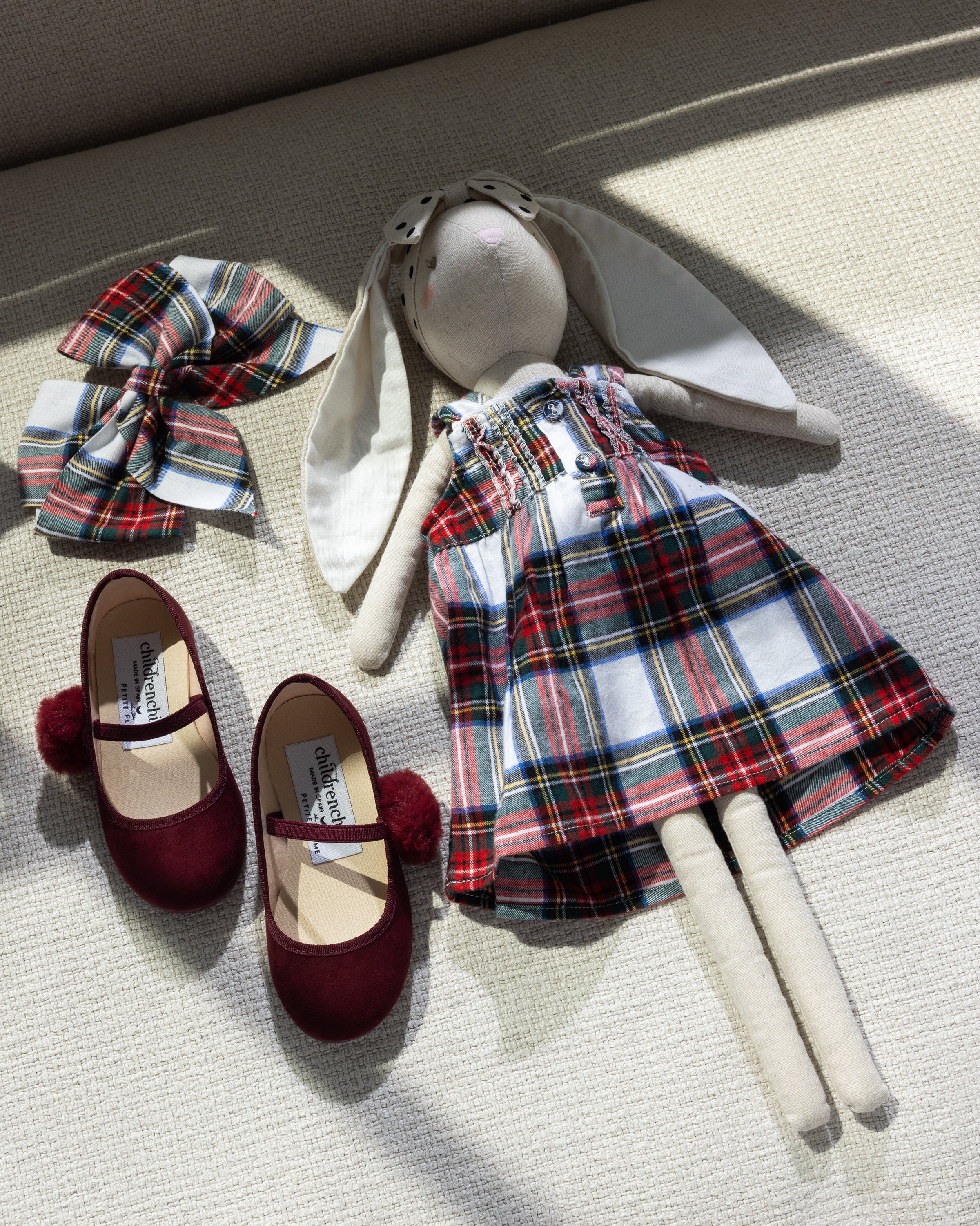 A fabric rabbit doll in a Balmoral Tartan plaid dress rests on a textured surface alongside Petite Plume Girls Hair Bows in Balmoral Tartan and red velvet ballet flats with pom-poms. Sunlight casts soft shadows, creating the perfect holiday gift display.