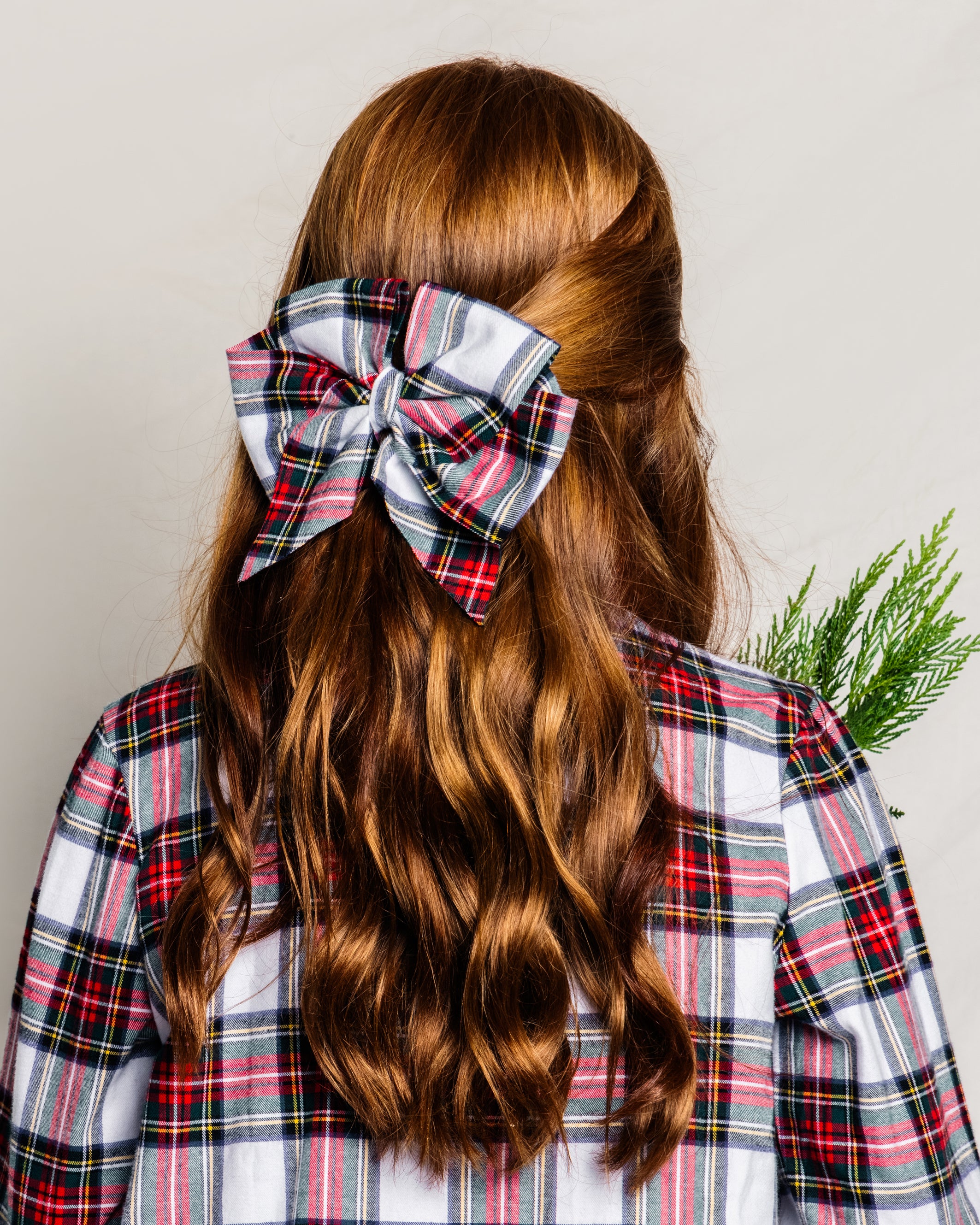 A person with long, wavy red hair, seen from behind in a plaid shirt, wears a large Petite Plume Balmoral Tartan hair bow. They hold a small bunch of green foliage, capturing the essence of girls holiday gifts.