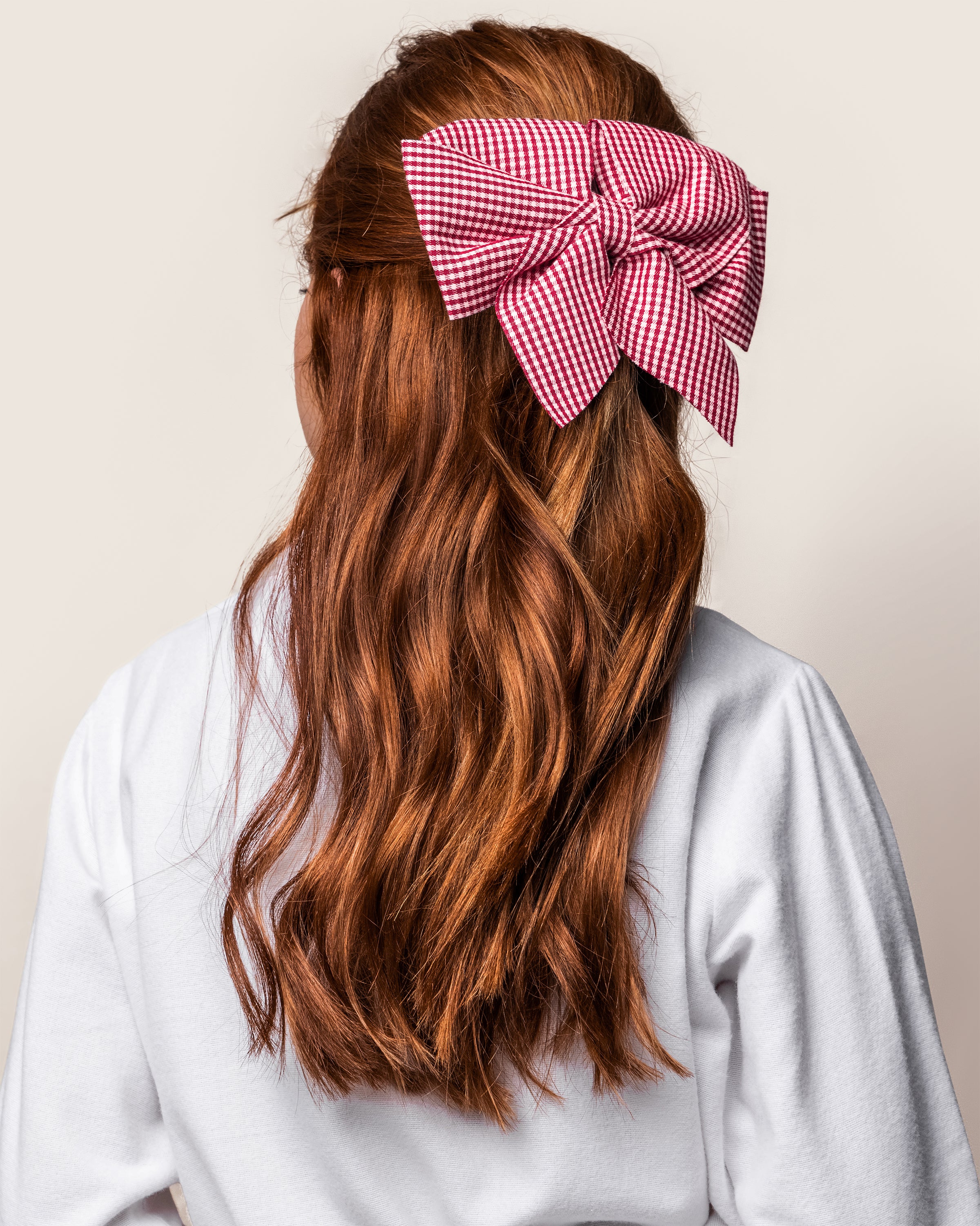 With long, wavy auburn hair, a person sports a large red and white Petite Plume Girls Hair Bow in Red Mini Gingham as an accessory. They wear a white top and face away from the camera against a light background.