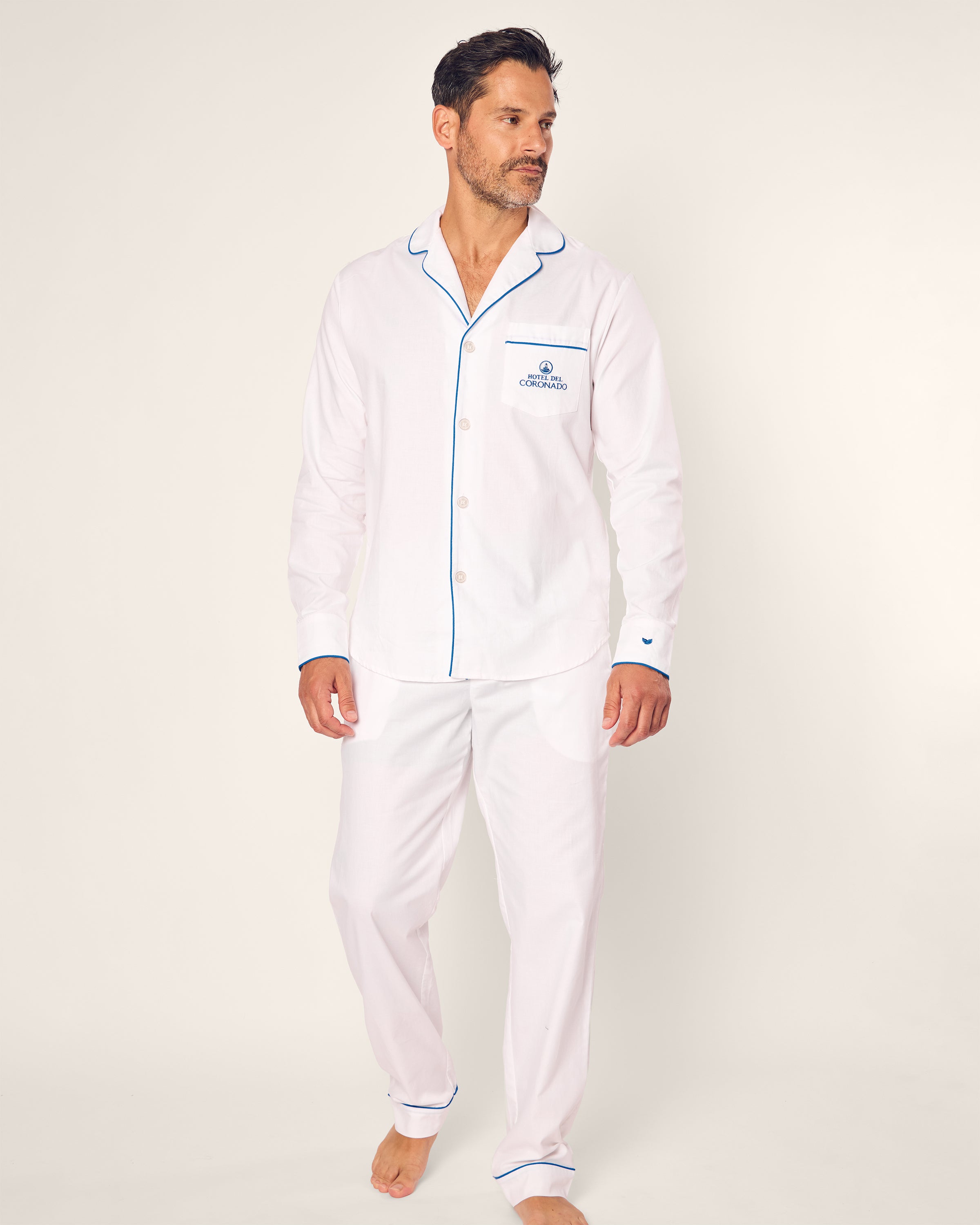 A man stands barefoot on a plain background wearing the Mens Twill Pajama Set from the Hotel del Coronado x Petite Plume collection, featuring luxurious white fabric with blue piping and embroidered text on the pocket. He appears relaxed in his casual posture.