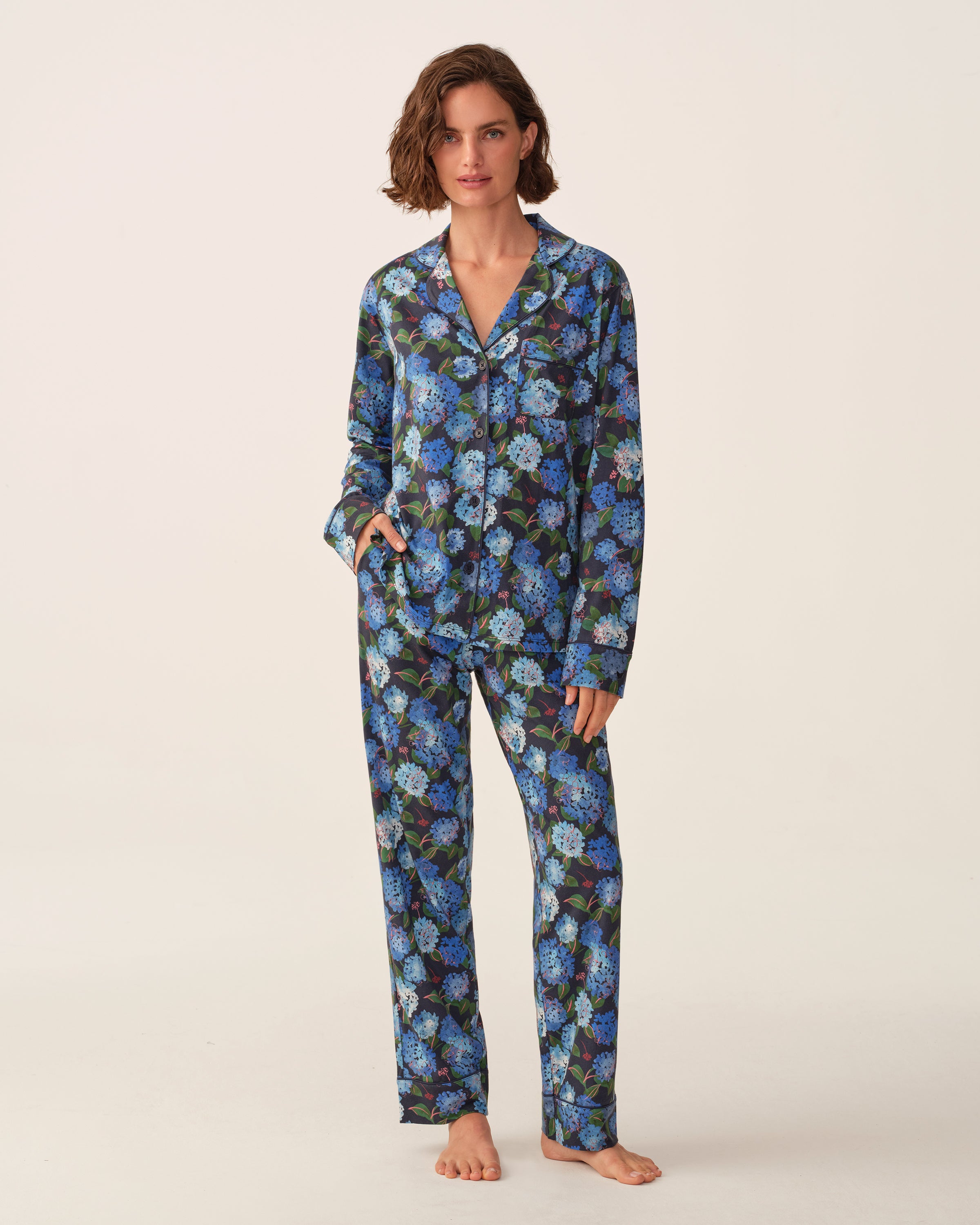 A person stands barefoot wearing the Petite Plume x Hedges Women’s Pima Pajama Set in Hydrangea, featuring a blue and purple floral pattern, posing against a plain light background.
