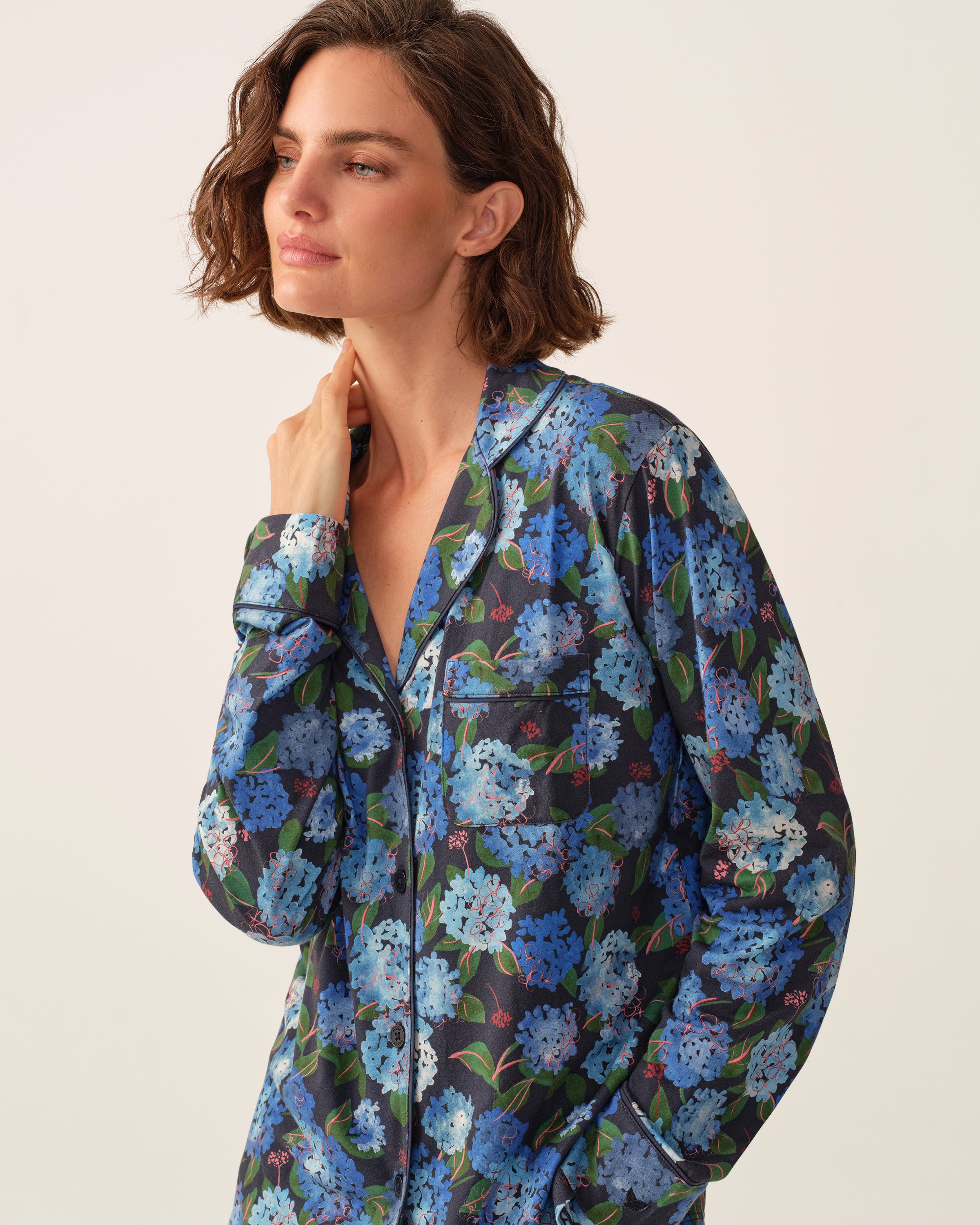 Woman with short brown hair wearing a dark pajama top with blue floral patterns, standing against a plain light background, looking to the side with a relaxed expression and her hand touching her neck.
