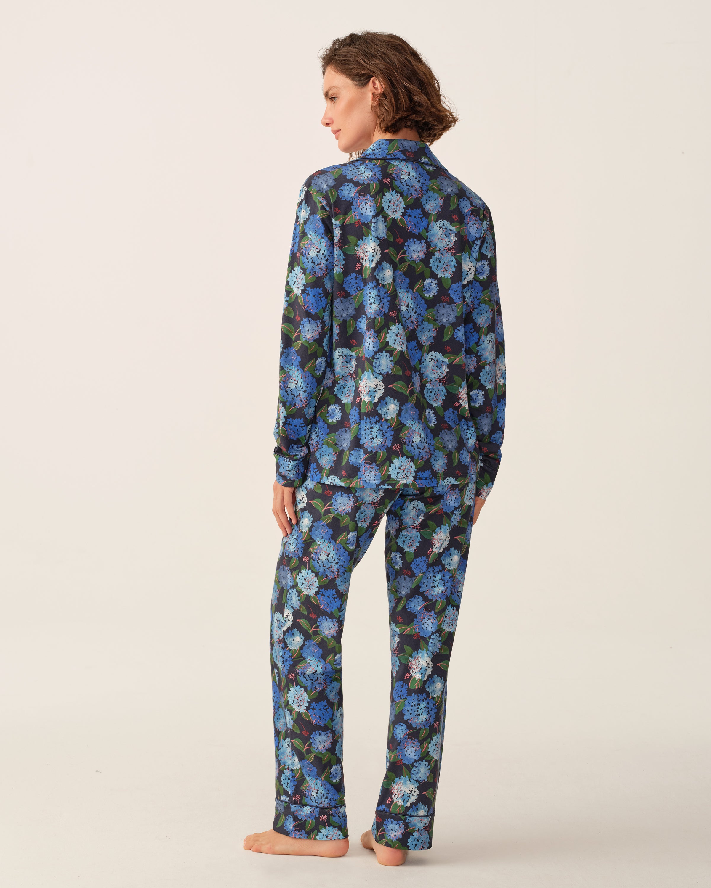 A person stands barefoot, facing away, wearing the Petite Plume x Hedges Women’s Pima Pajama Set in Hydrangea—featuring a blue and purple floral pattern on a dark background—against a plain, light backdrop.