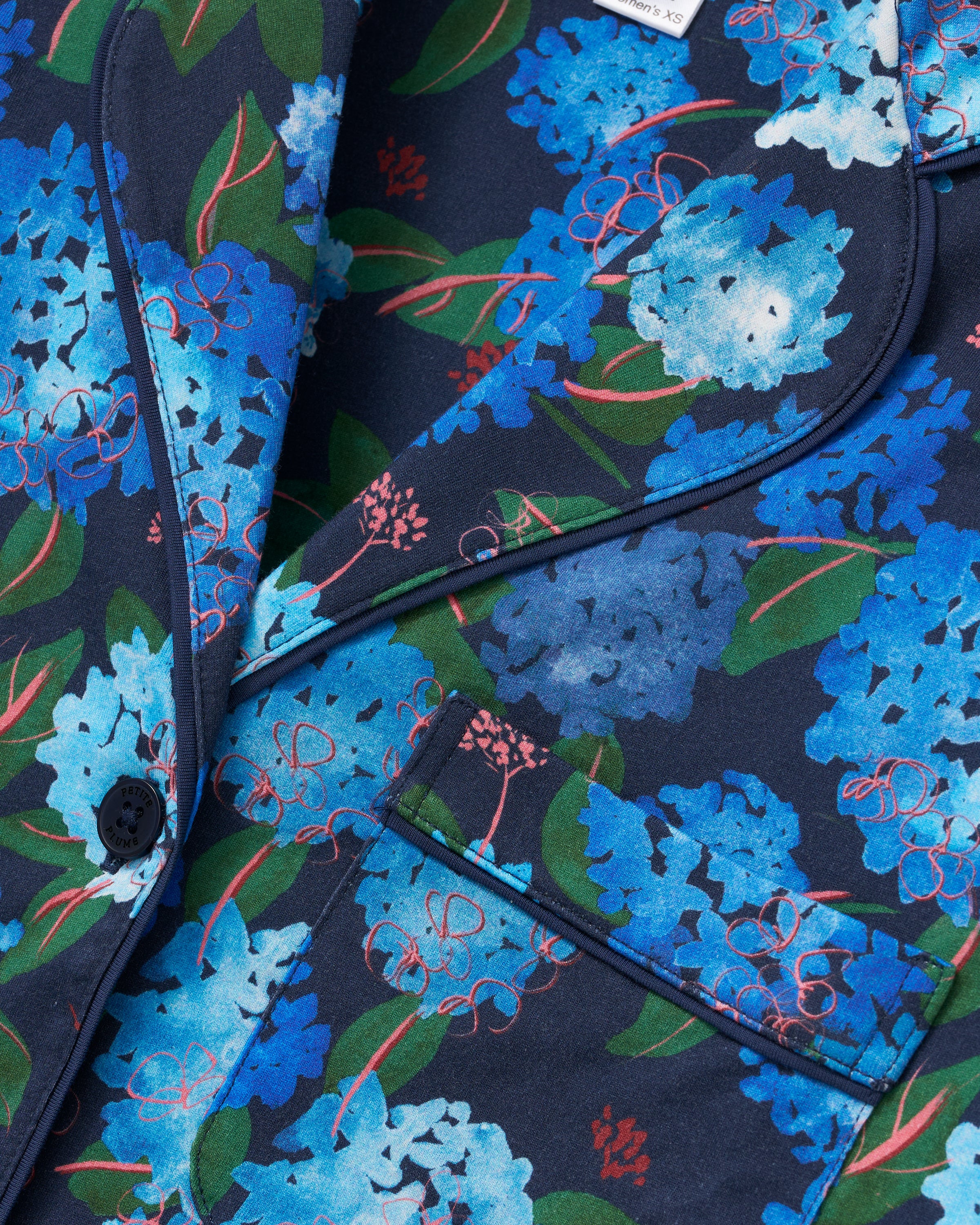 Close-up of the Petite Plume x Hedges Women’s Pima Pajama Set in Hydrangea, showcasing a navy blue fabric adorned with a vibrant blue floral pattern, accented by green leaves and red branches. The pajama set features detailed piping, a front pocket, and a black button.