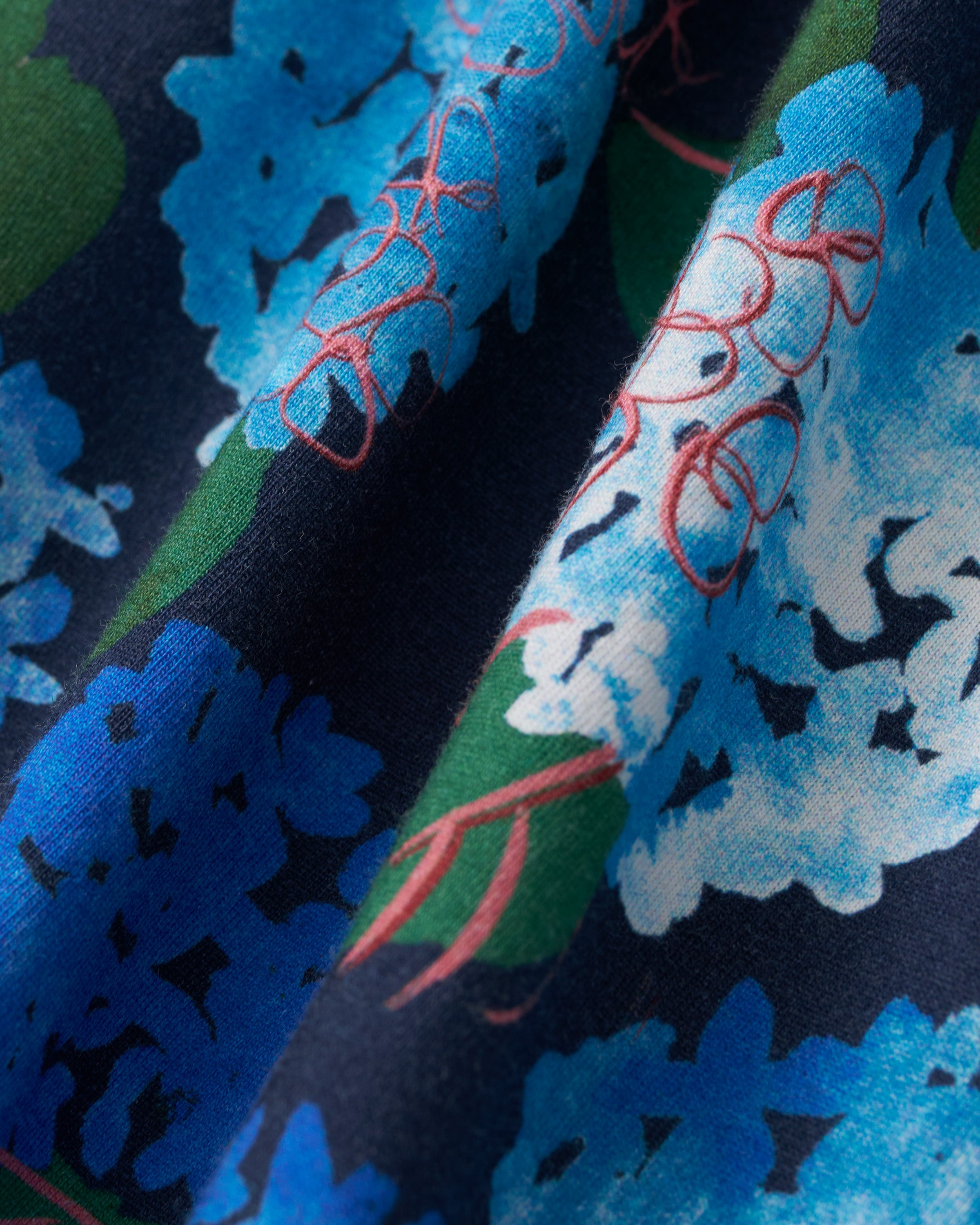 Close-up of the Petite Plume x Hedges Women’s Pima Pajama Set in Hydrangea, featuring fabric with a floral pattern in shades of blue and white, set against a dark blue background, accented with green leaves and pink flower outlines. The soft pima cotton drapes in gentle folds.