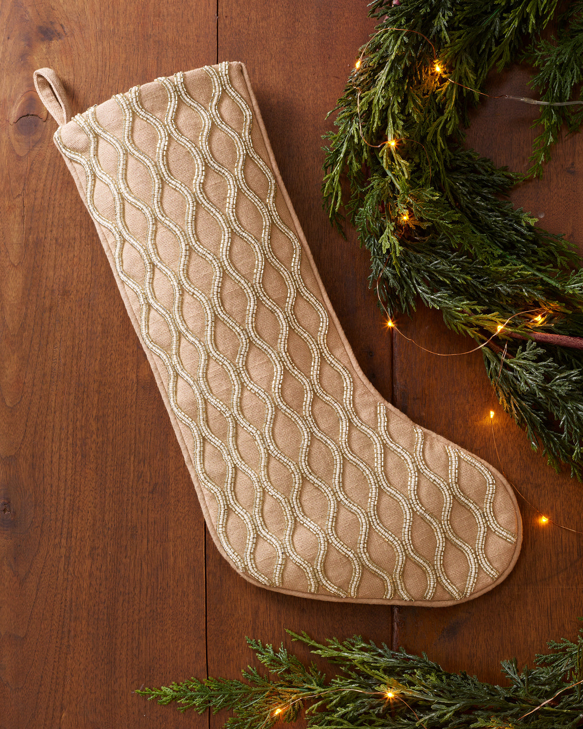 Beaded Christmas Stocking in Neutral made of tan fabric with wavy lines of silver beads, resting on a wooden surface next to an evergreen garland with string lights.