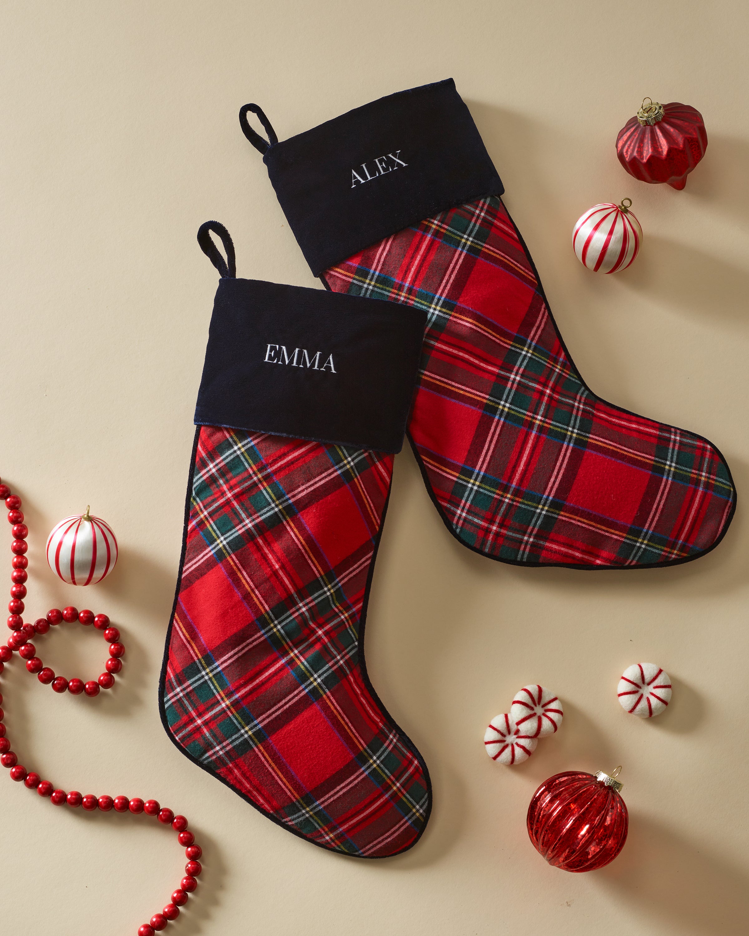 Two red plaid Christmas Stockings in Imperial Tartan by Petite Plume, with plush velour cuffs labeled Emma and Alex, are adorned with red and white ornaments—striped balls, candy swirls—and a string of red beads on a beige surface.