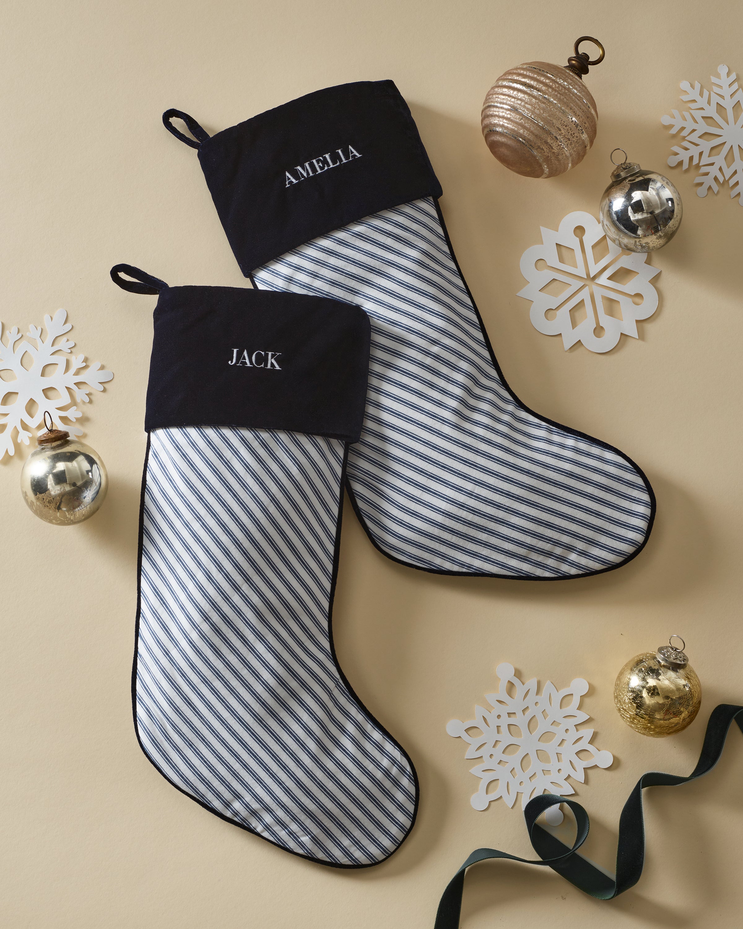 Two Petite Plume Christmas stockings in Navy French Ticking, with plush velour cuffs labeled AMELIA and JACK, are surrounded by gold and silver ornaments on a beige background. Perfect monogrammable holiday stockings to personalize your festive décor.