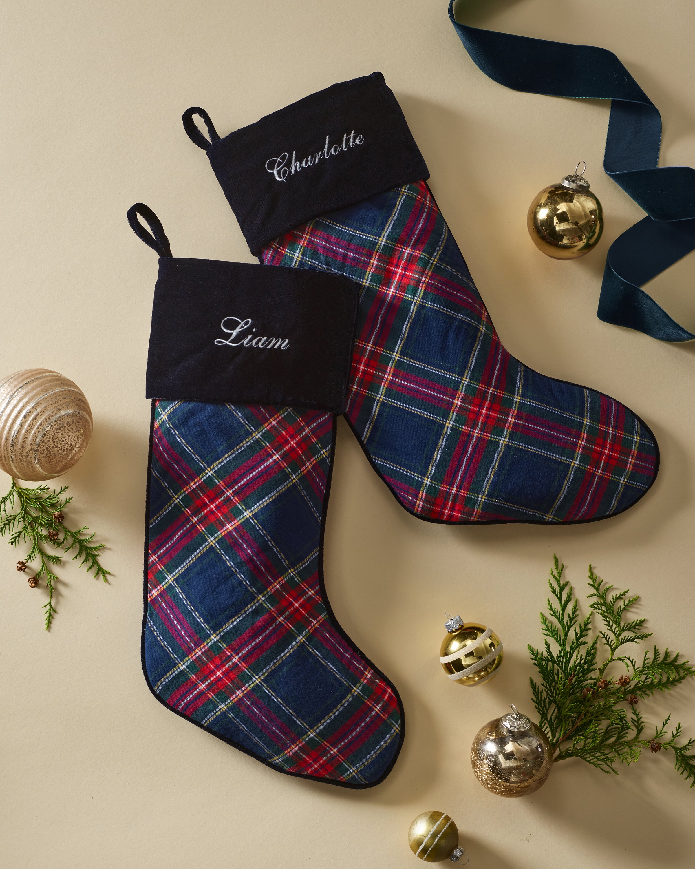 Two Windsor Tartan Christmas Stockings by Petite Plume, bearing Charlotte and Liam, lie flat. Monogrammed and elegant, surrounded by gold ornaments, satin ribbons, and evergreen branches on a light background, they make the perfect holiday display.