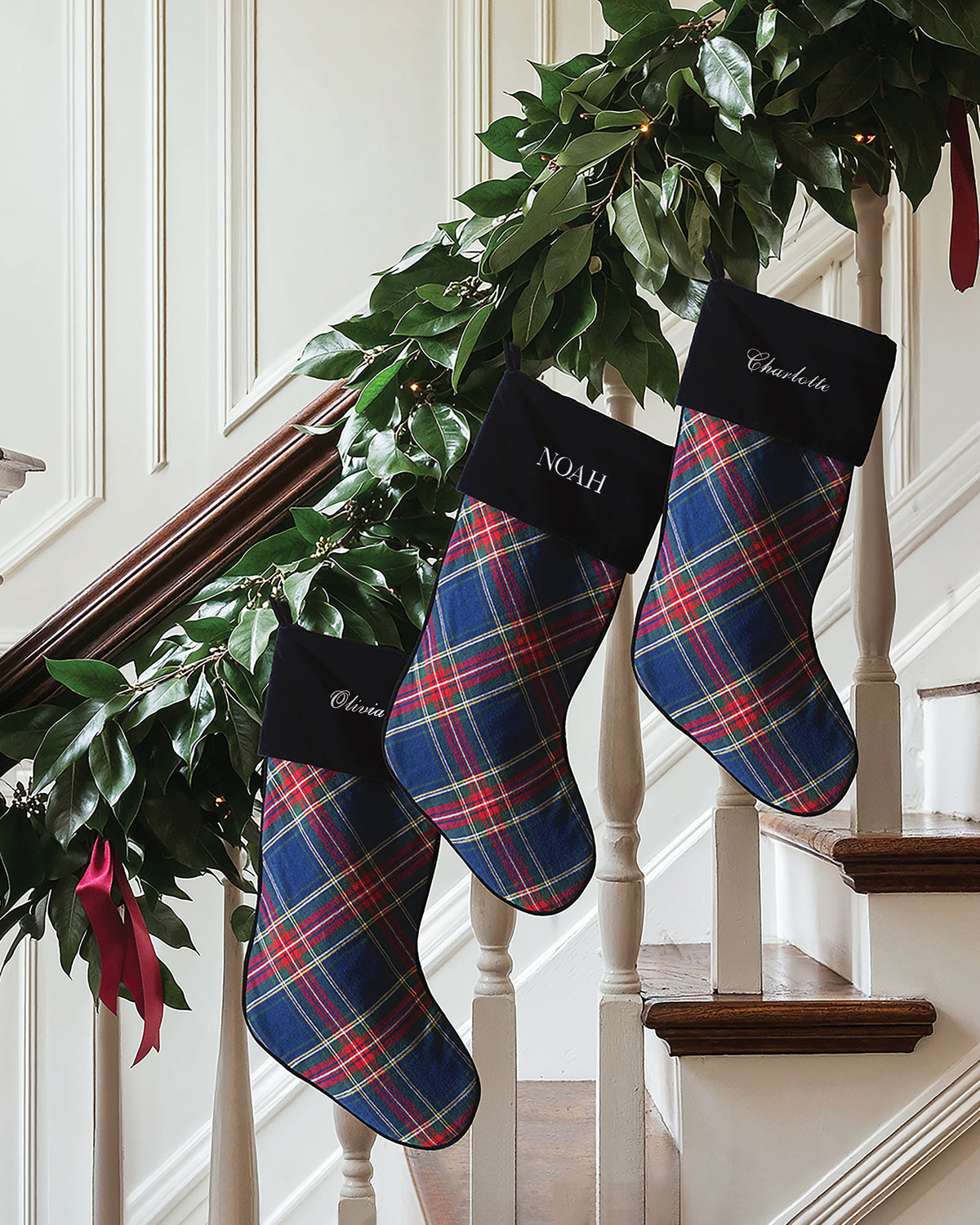 Three Christmas Stocking in Windsor Tartan stockings with blue, red, and green tartan patterns hanging from a staircase banister decorated with greenery.