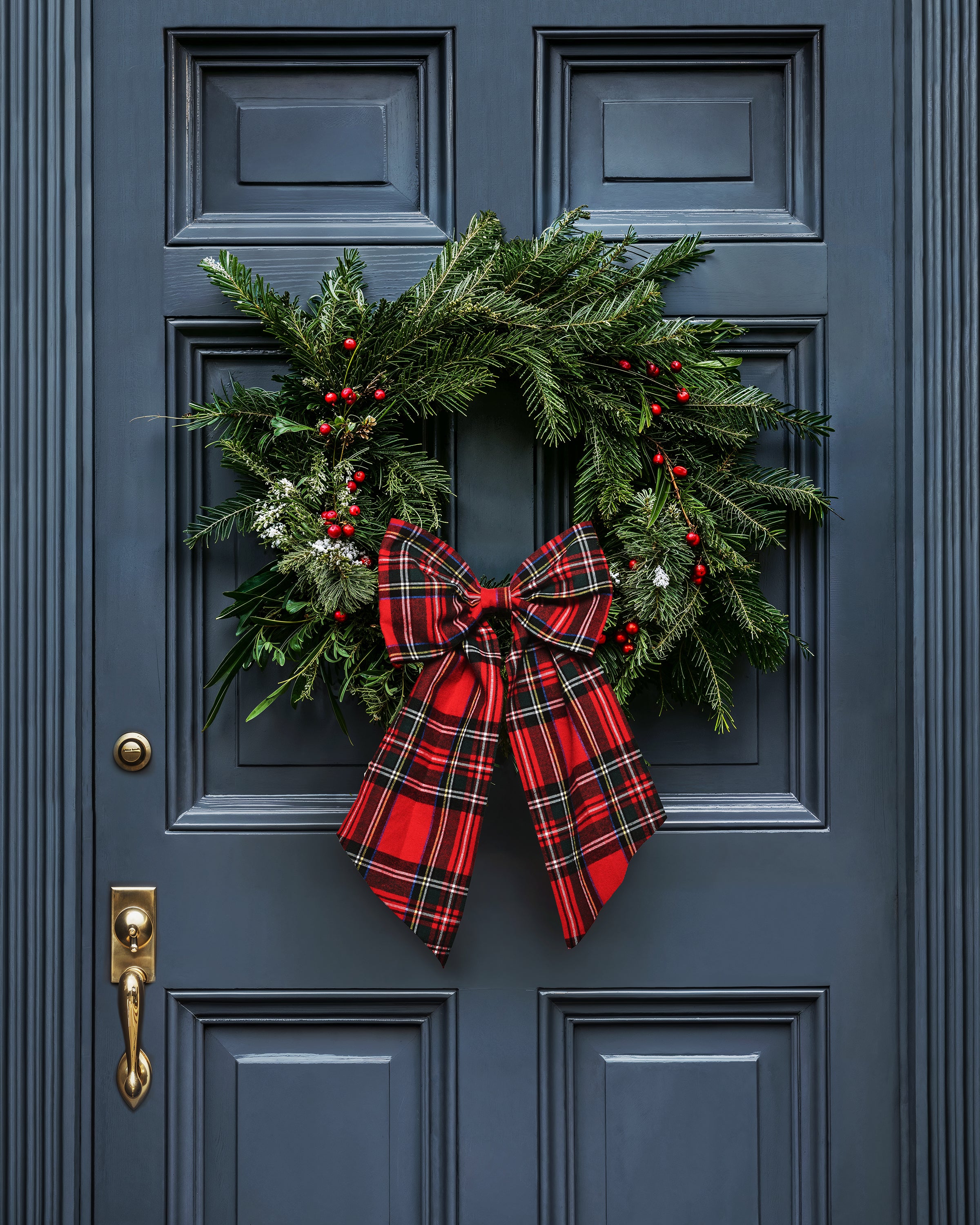 A Petite Plume Wreath Bow in Imperial Tartan with green pine, red berries, and white accents adorns the dark blue door. This holiday decor is highlighted by a striking red and black plaid bow with a brass handle and knocker for elegance.