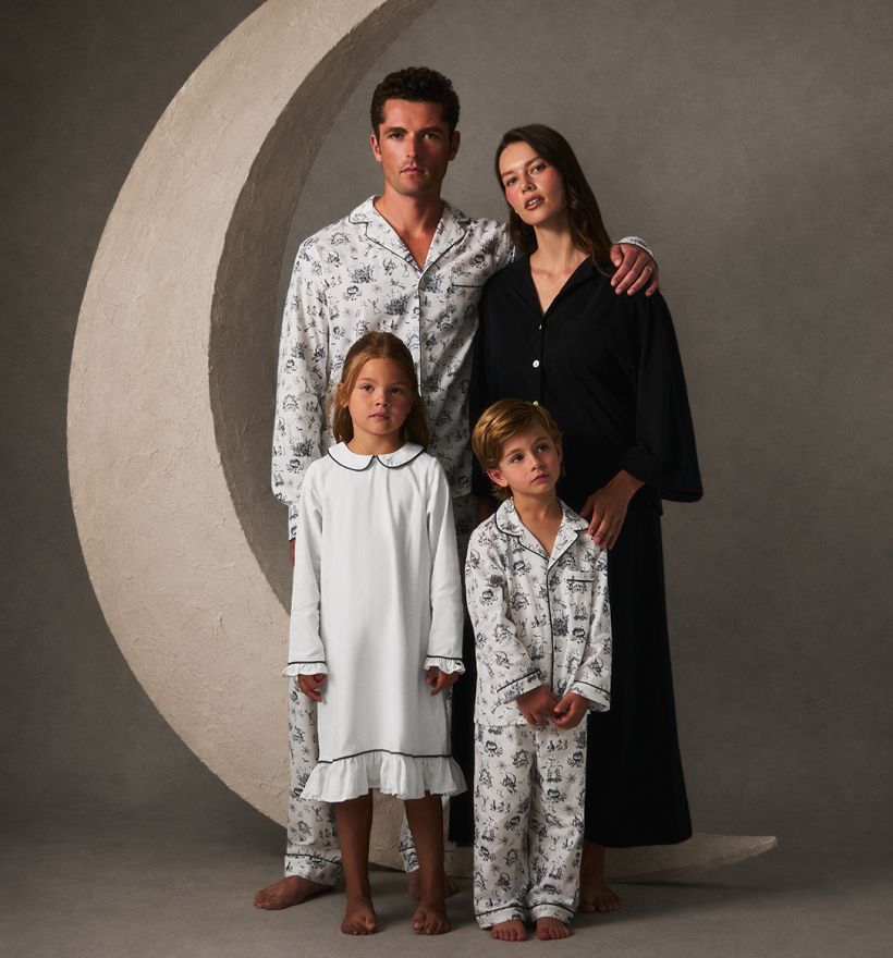 A family of four, all in pajamas, poses barefoot in front of a large crescent moon prop and a neutral backdrop. The adults stand behind the children, with relaxed, calm expressions.