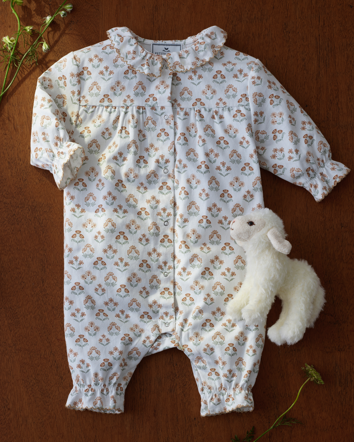 Baby’s Twill Madeline Romper in Estate Garden, featuring a white floral pattern with a ruffled collar and cuffs, is displayed flat on a wooden surface alongside a small white plush lamb toy and several sprigs of greenery.