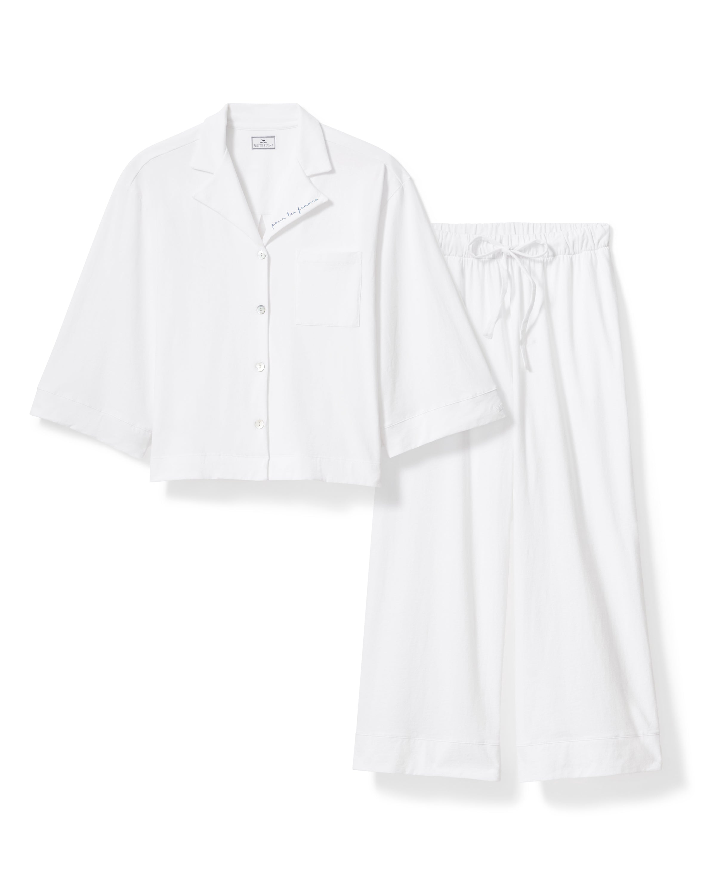 A woman with long, wavy dark hair models The Olivia Wide Leg Pima Pajama Set With "Pour Les Femmes" Embroidery, featuring a loose white button-up shirt with collar and front pocket, gazing slightly upward against a plain background.