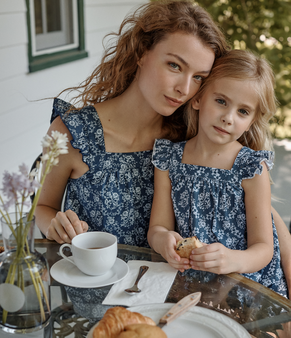 A woman and a young girl in matching blue floral dresses sit closely at an outdoor table with a cup of coffee, pastries, and flowers in a vase.