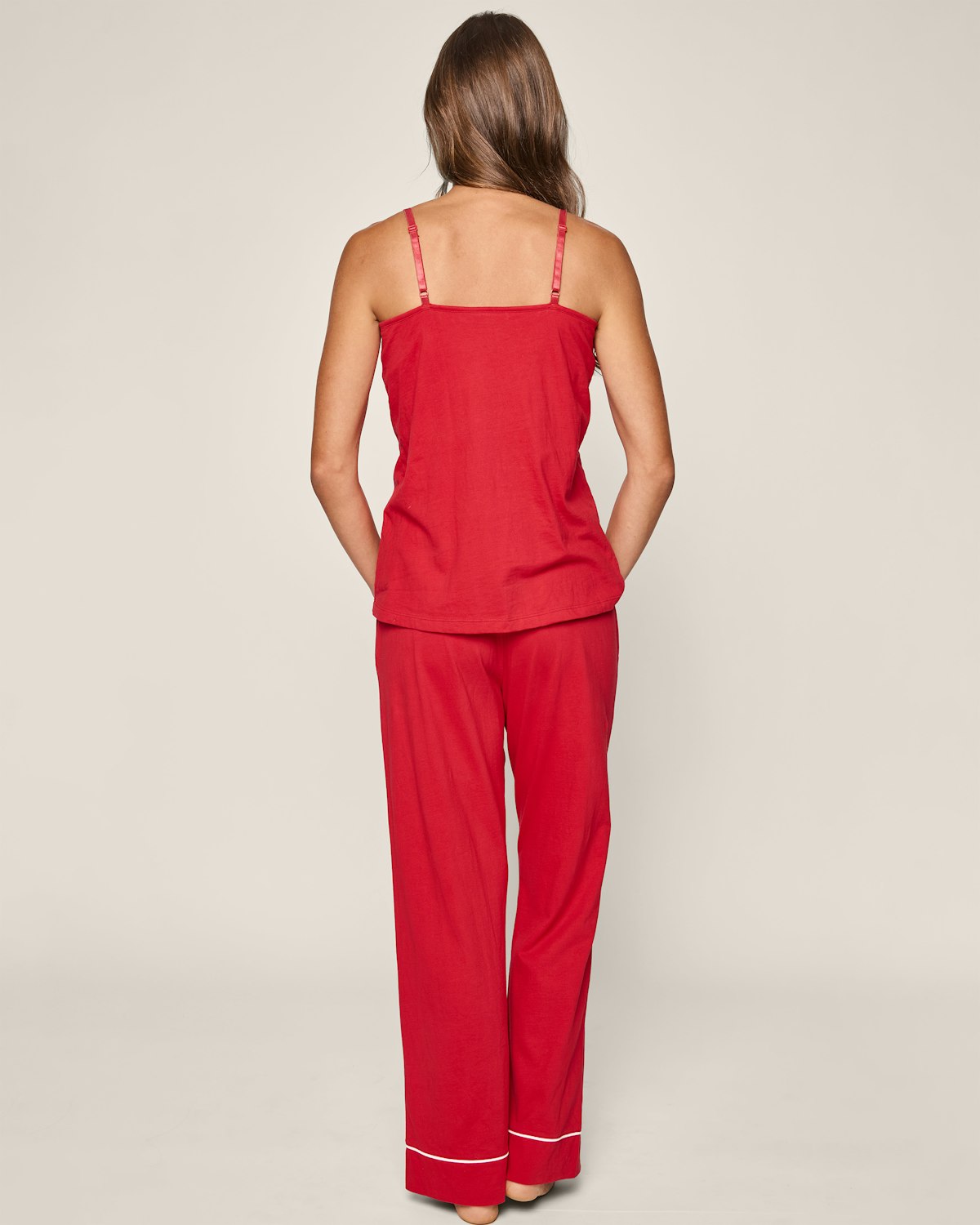 A woman with long brown hair stands facing away in a red pajama set made of luxurious Pima cotton by Petite Plume. The sleeveless top and pants, from the Womens Pima Maternity Pants collection, feature elegant white piping at the hem against an off-white background.