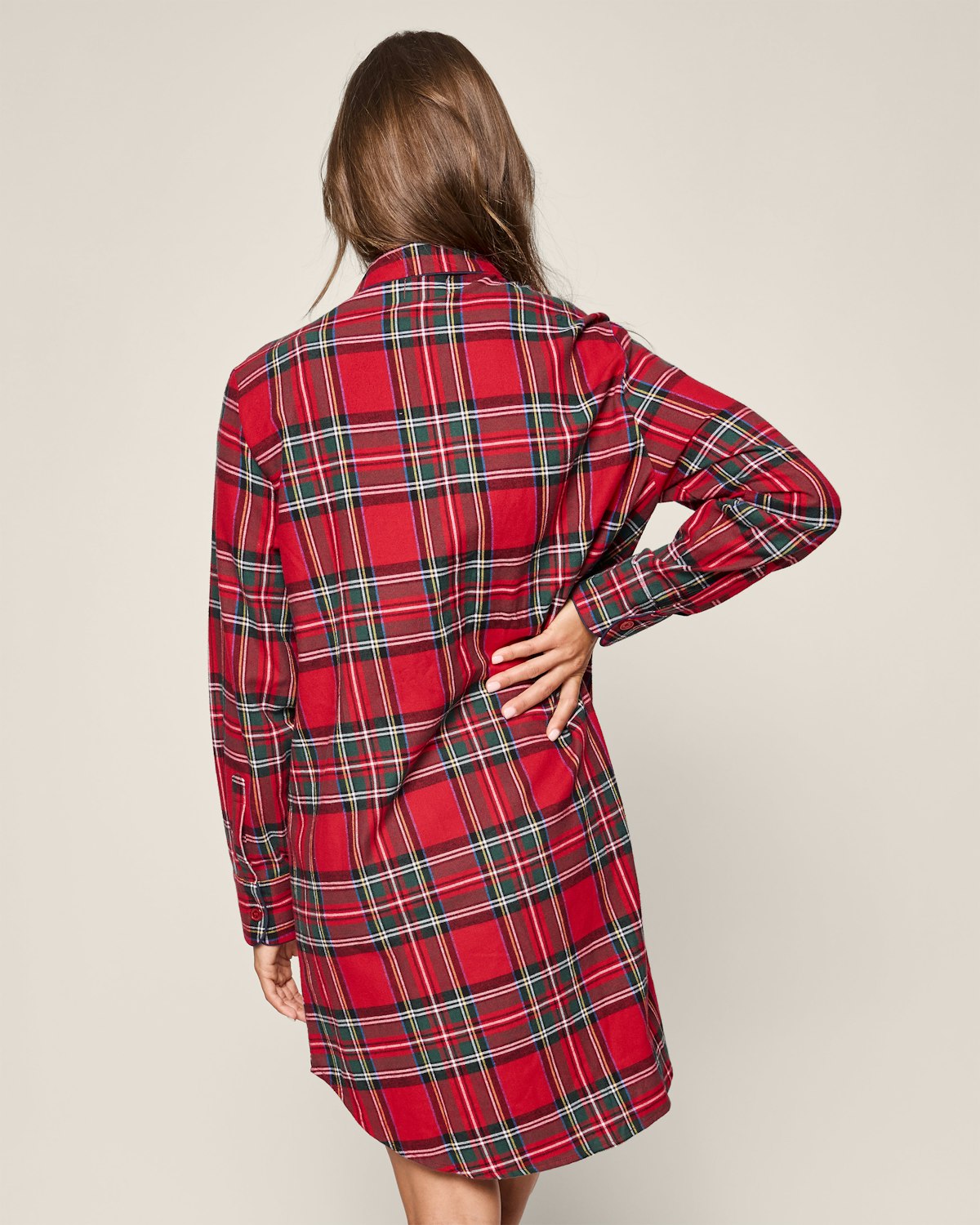 A person with long brown hair in a red Imperial Tartan maternity nightshirt by Petite Plume stands with their back to the camera, one hand on their hip, against an off-white background.