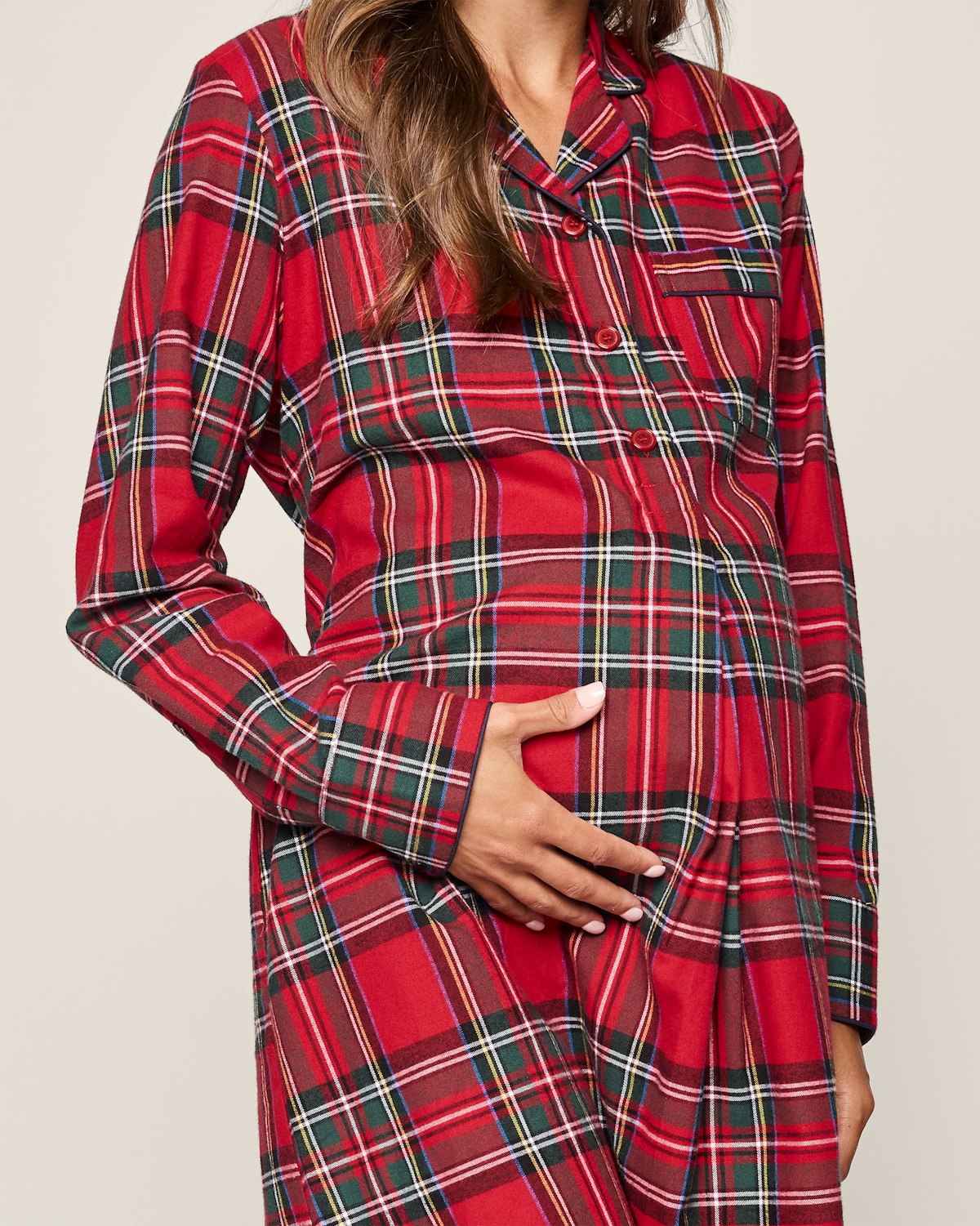 Dressed in Petite Plumes Womens Brushed Cotton Maternity Nightshirt in Imperial Tartan, a person gently cradles their pregnant belly. The plain background emphasizes the luxurious red and green Scottish plaid of the fabric.