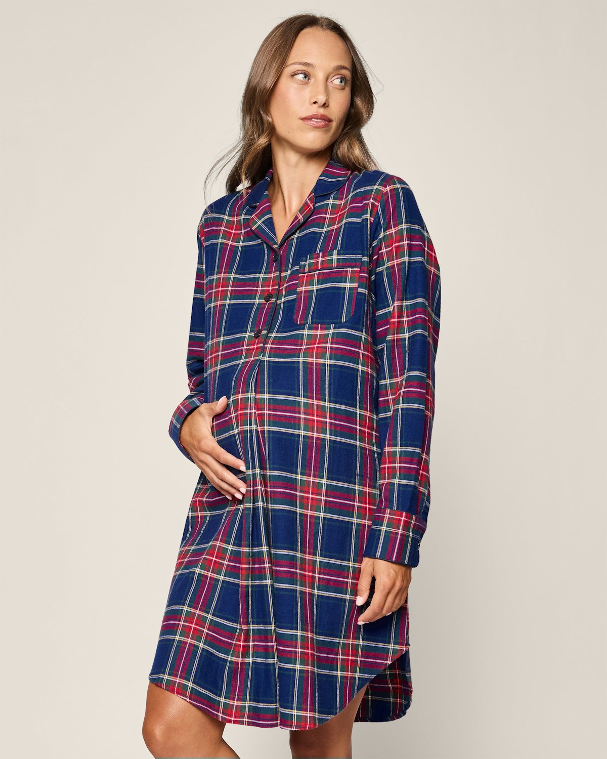 A person in Petite Plume’s Womens Brushed Cotton Maternity Nightshirt in Windsor Tartan, featuring a blue, red, and white plaid pattern, stands against a plain backdrop. With long brown hair and side gaze, they touch their abdomen.