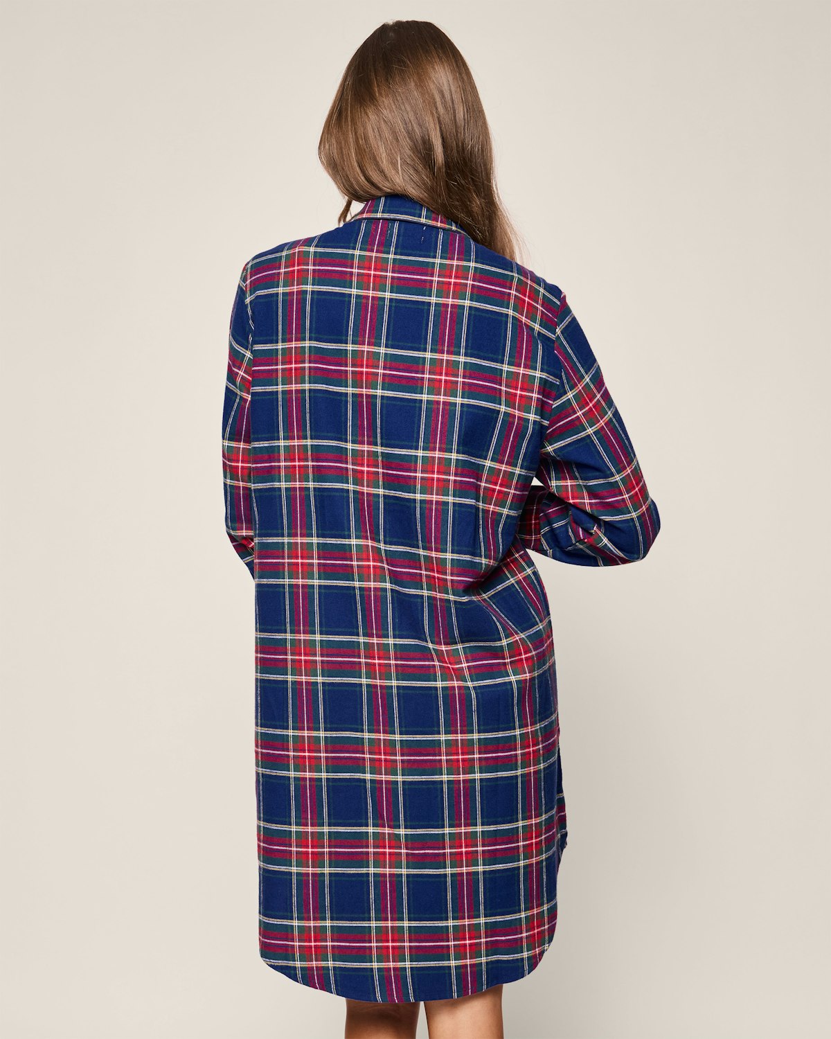 A woman with long brown hair faces away, wearing Petite Plumes Womens Brushed Cotton Maternity Nightshirt in Windsor Tartan. The nightshirt extends to just above her knees, offering a relaxed fit and featuring blue, red, and green plaid against a plain background.