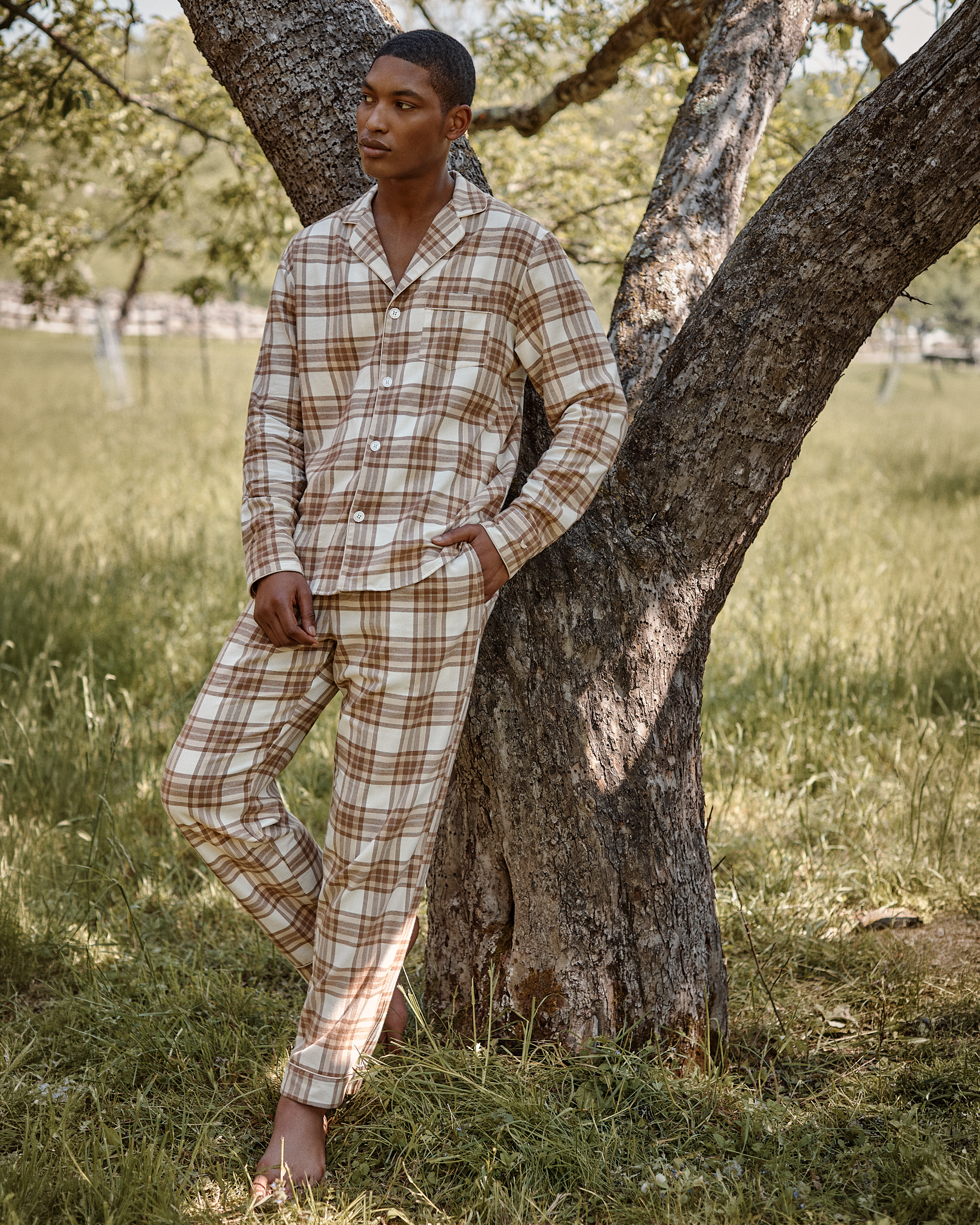 A person wearing the Men’s Twill Pajama Set in Cotswolds Tartan leans against a tree in a grassy outdoor setting on a sunny day.