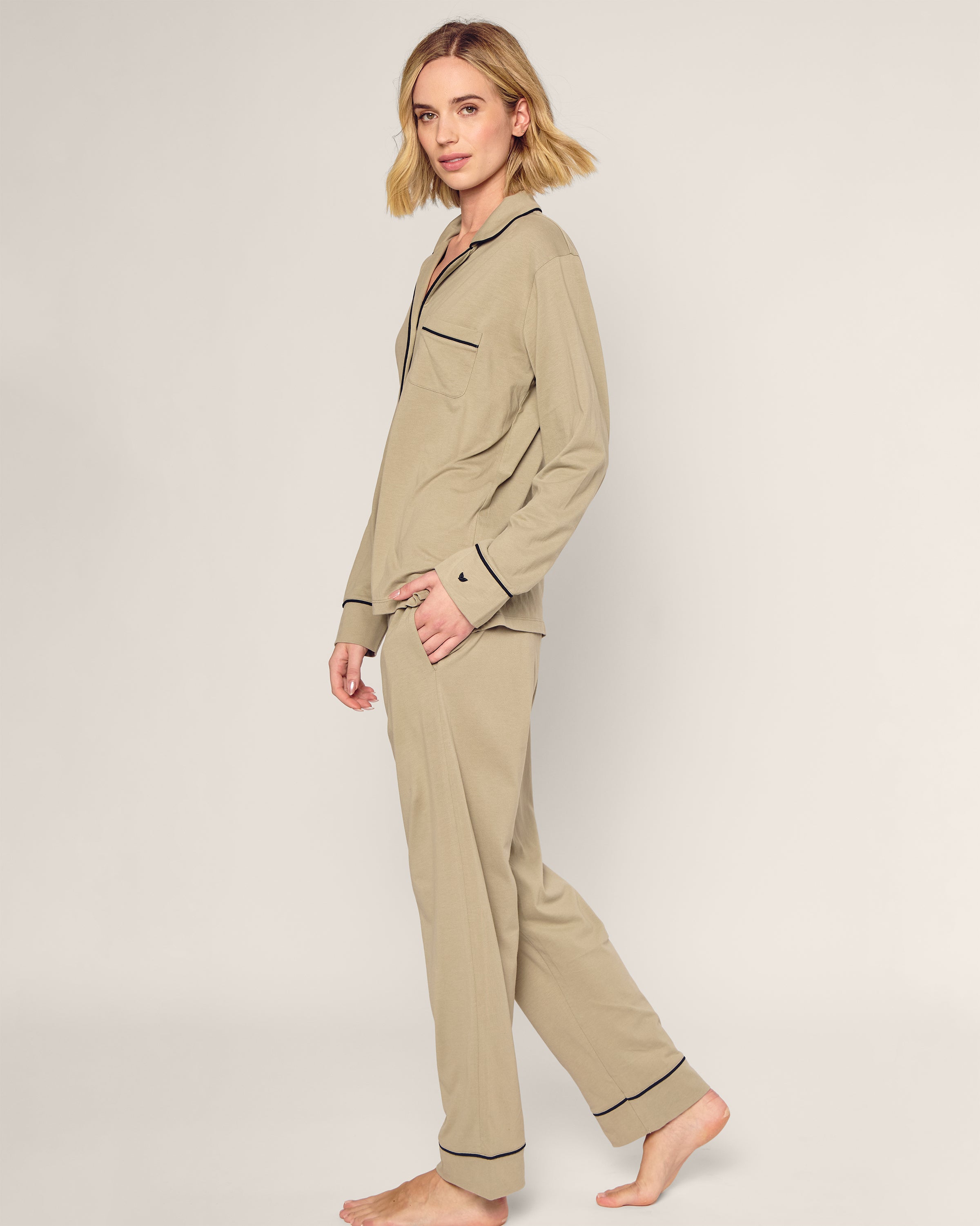 A person with shoulder-length blonde hair is wearing the Petite Plume x Parachute Womens Pima Pajama Set in Moss. They are barefoot against a plain background, slightly turned to their left, with a relaxed expression.