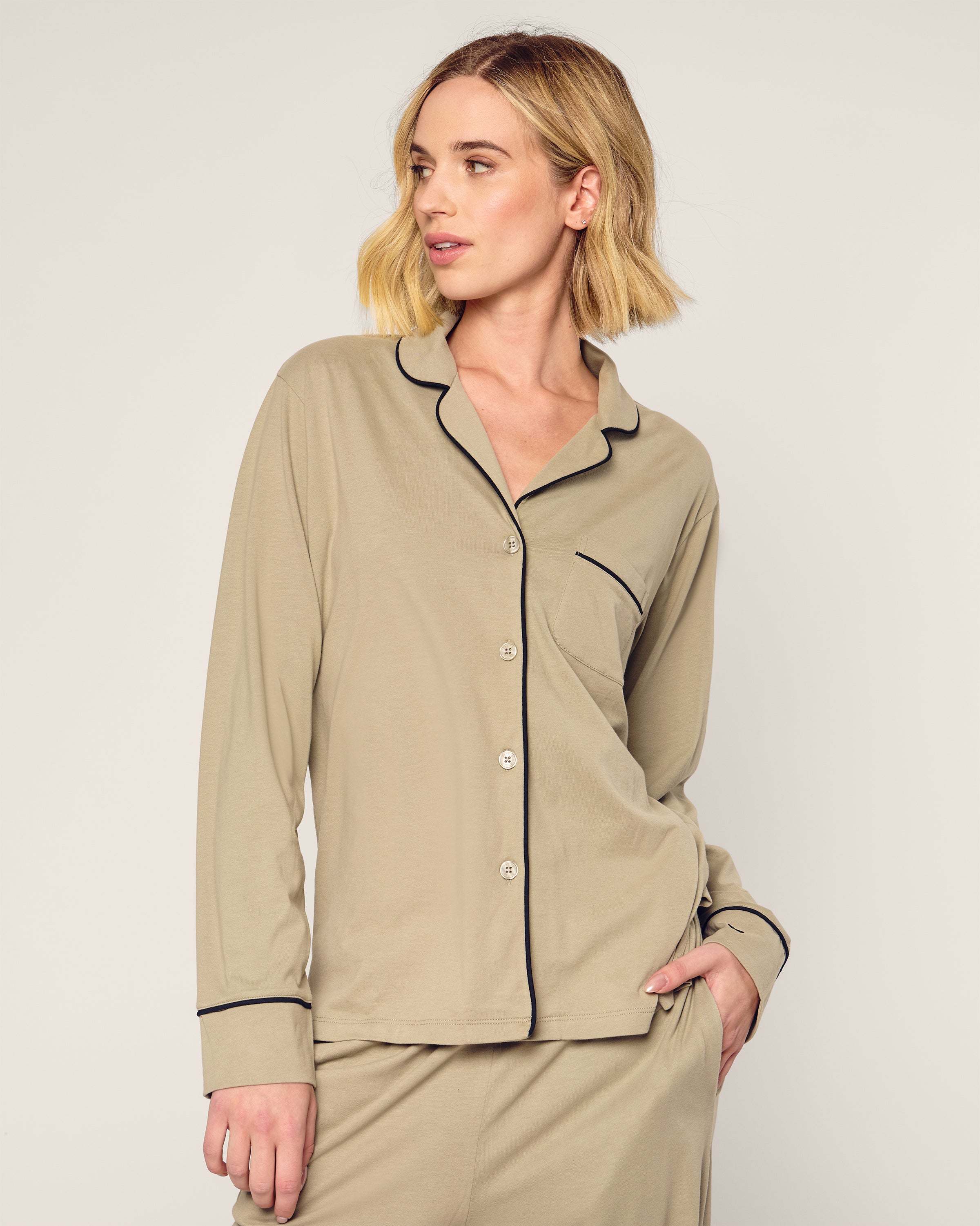 A person with shoulder-length blonde hair wears the Petite Plume x Parachute Womens Pima Pajama Set in Moss. The buttery soft set features a button-up top with black trim and a breast pocket. They stand relaxed against a neutral backdrop, gazing sideways with one hand in their pocket.