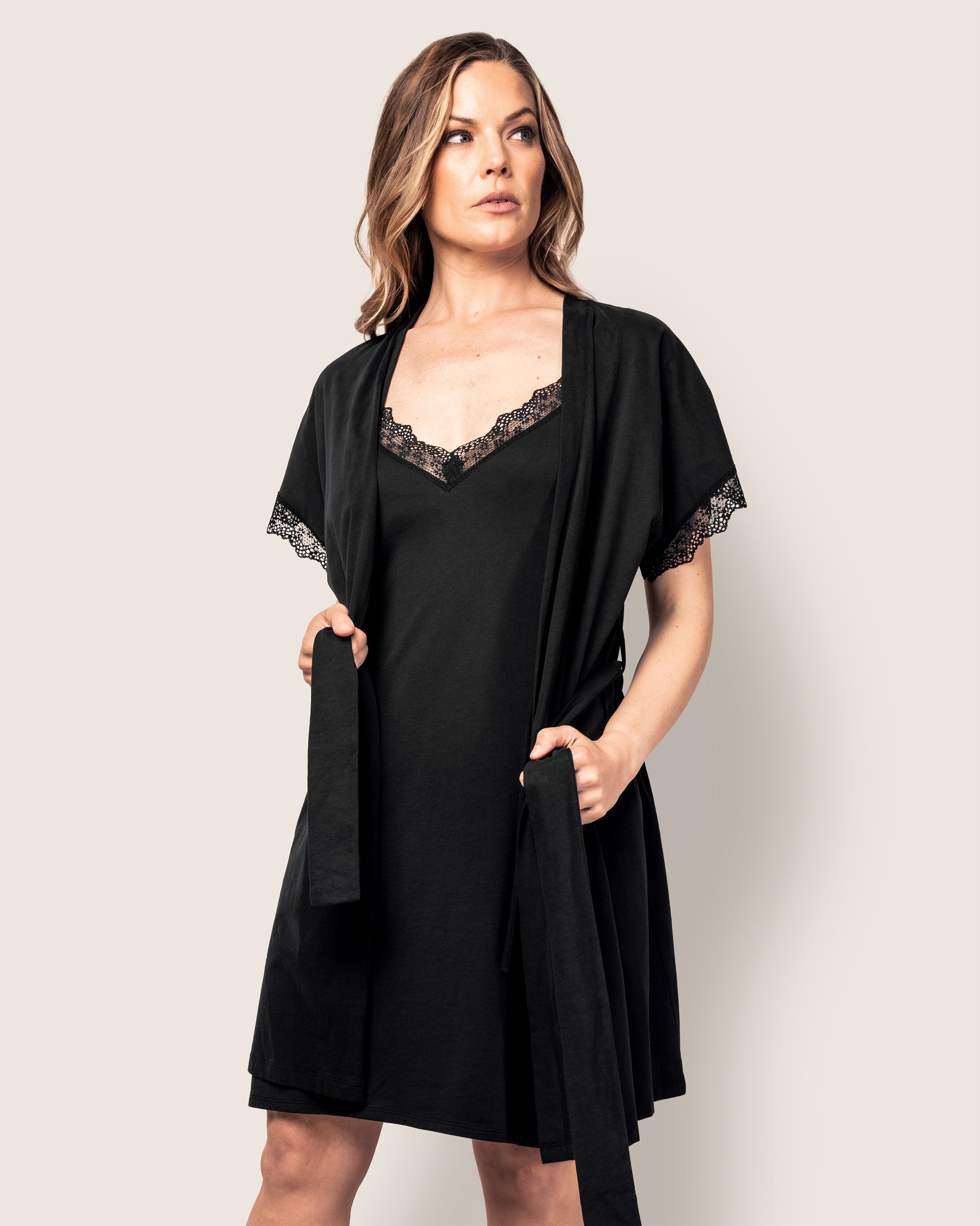 A woman wears Petite Plumes Womens Pima Nightgown with Lace in Black, featuring lace detailing at the neckline and sleeves. She appears focused and confident, with loose waves in her hair against an off-white background.