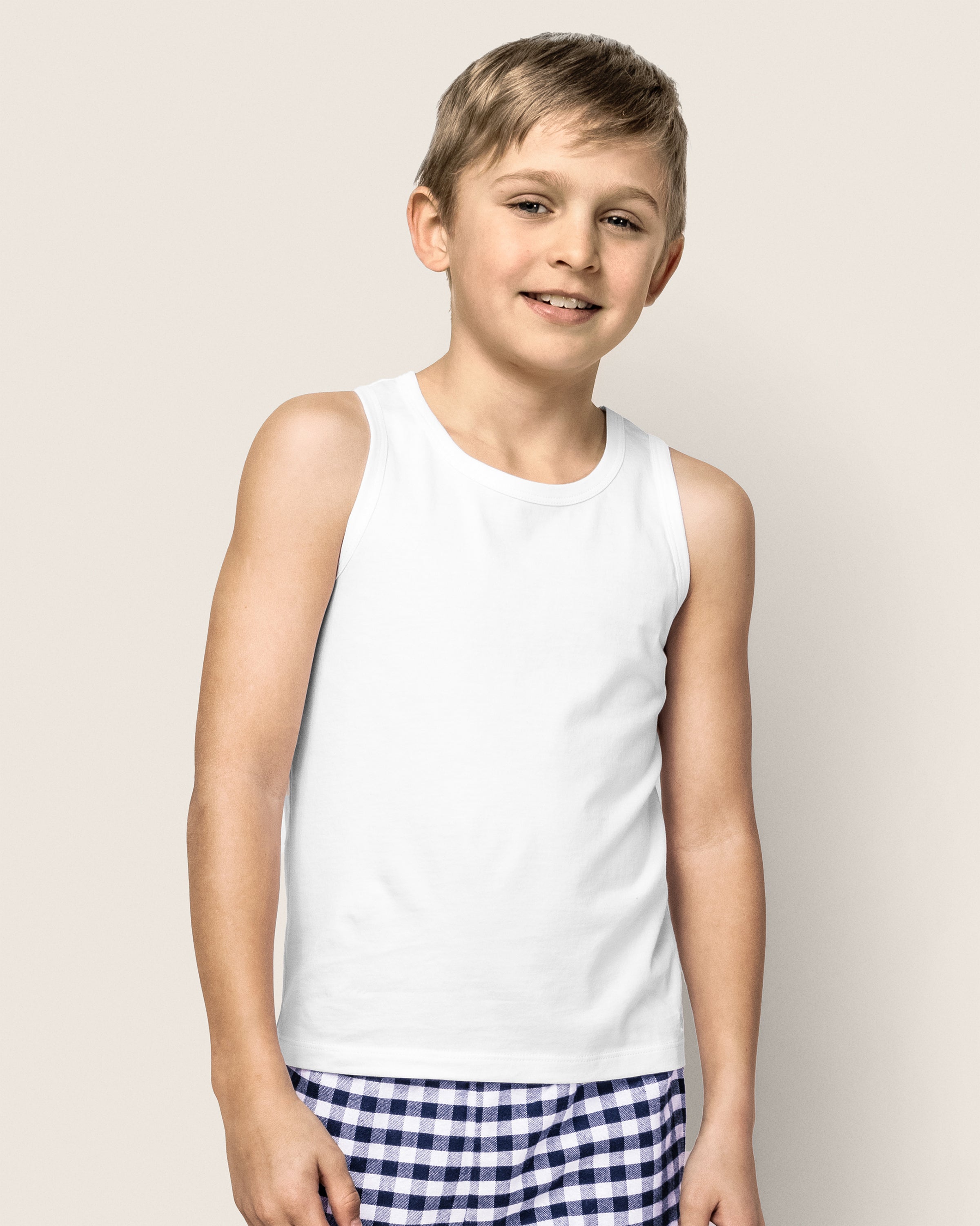 A young blonde boy smiles at the camera, wearing a comfy set featuring Petite Plumes Kids Pima Tank Top in White and blue and white checkered shorts. The scene is set against a light-colored plain background.