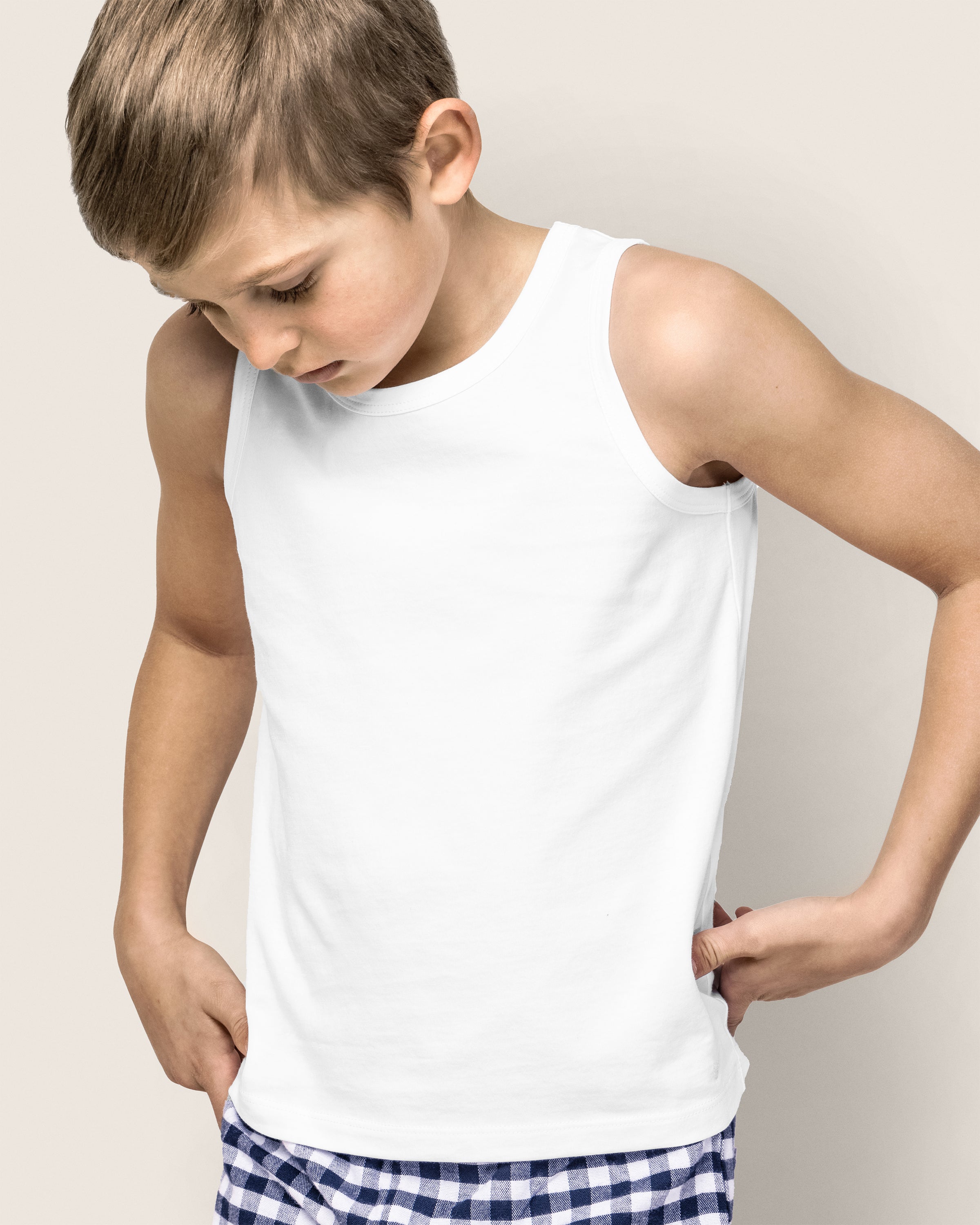 A young boy in a Petite Plume Kids Pima Tank Top in White and blue checkered shorts, made from soft Peruvian Pima cotton, looks down with his hands on his hips against a plain background.