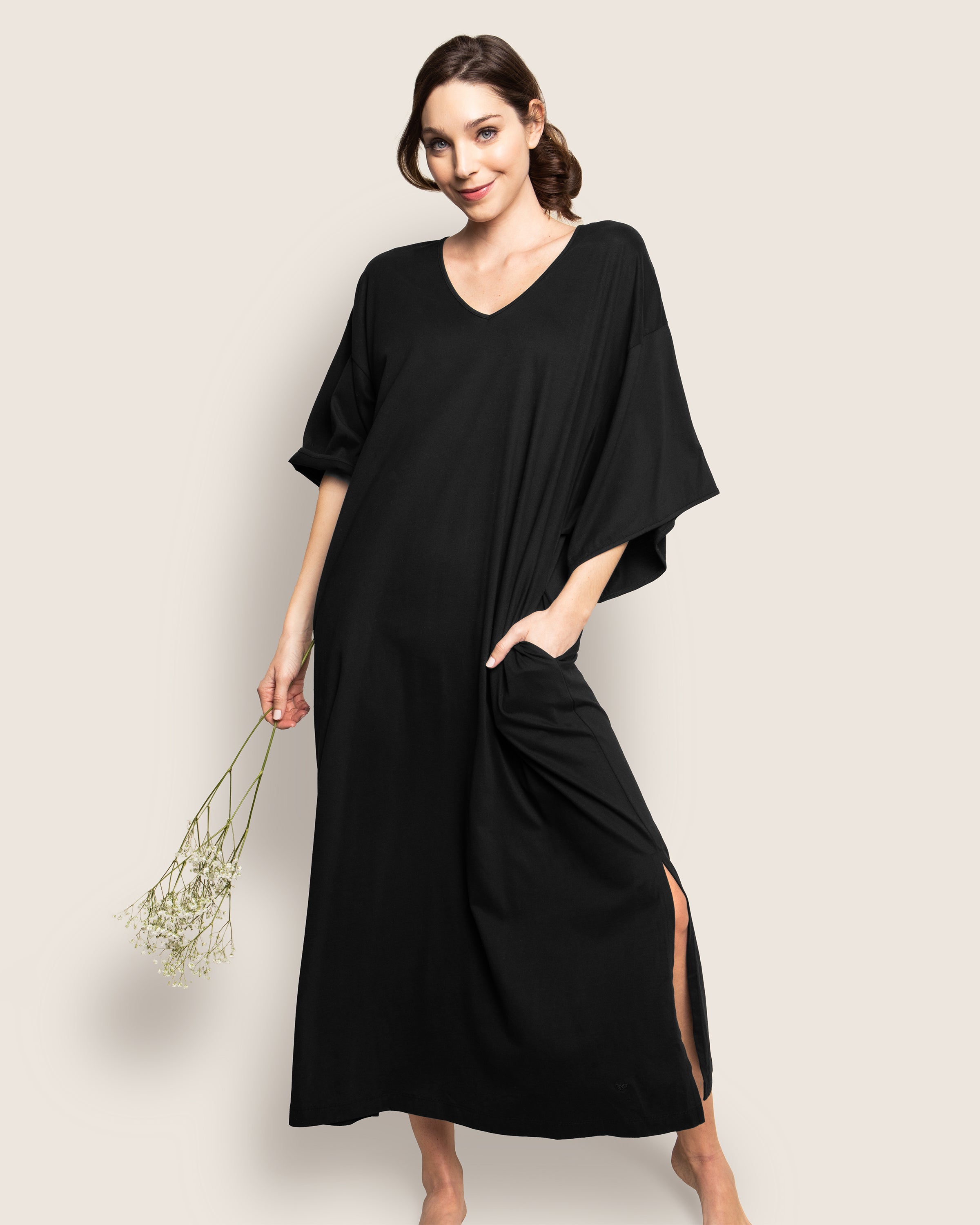 A person in a Womens Pima Caftan by Petite Plume, crafted from 100% Peruvian Pima cotton jersey knit, stands with a small bouquet of white flowers. The loose-fitting black dress has short sleeves, a V-neckline, side slits, and pockets against a plain light background.