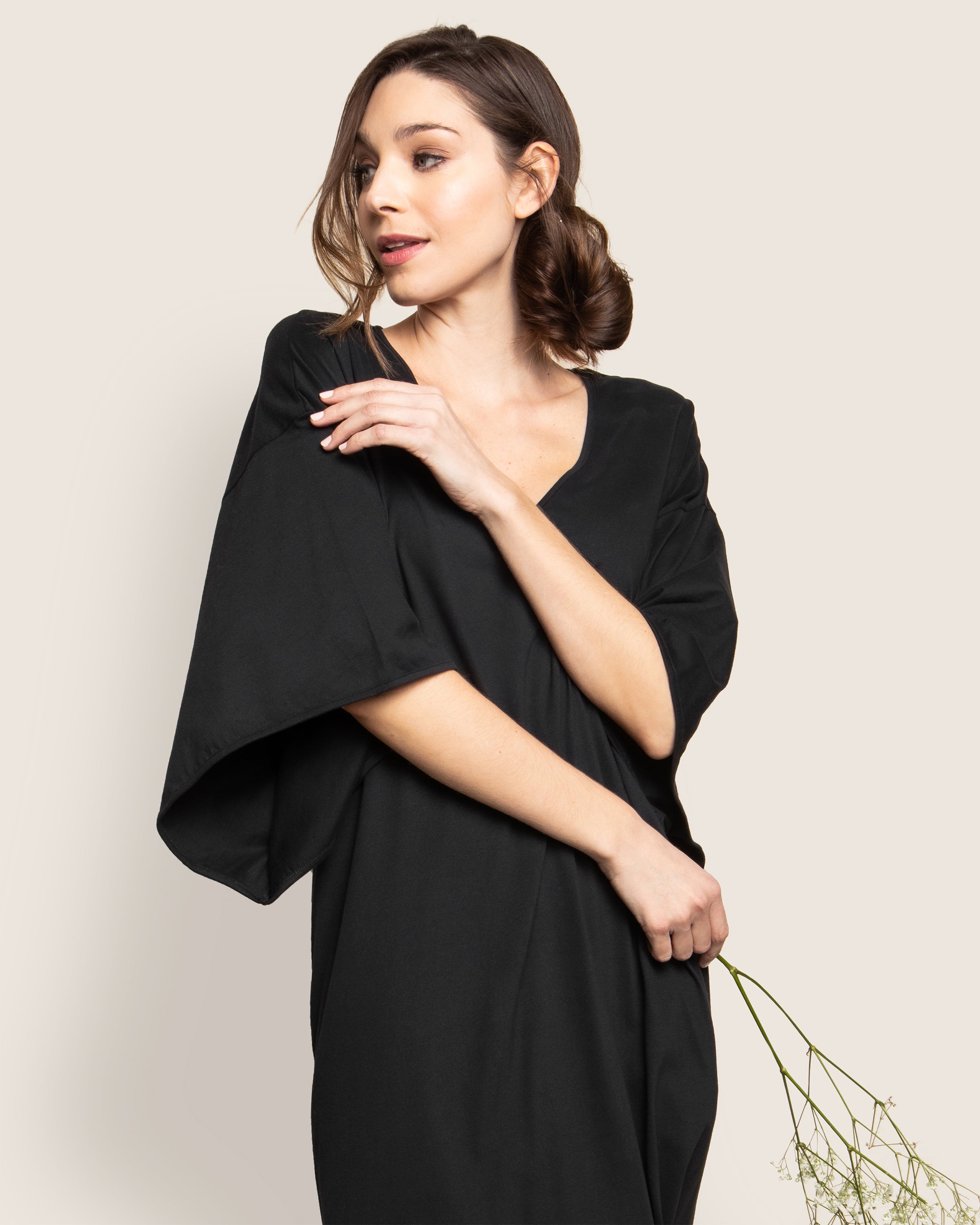 The woman wears a Petite Plume Womens Pima Caftan in Black, made from 100% Peruvian Pima cotton, and holds delicate flowers. Her hair is styled in a low bun as she stands against a plain cream backdrop, gazing slightly to the side with a serene expression.