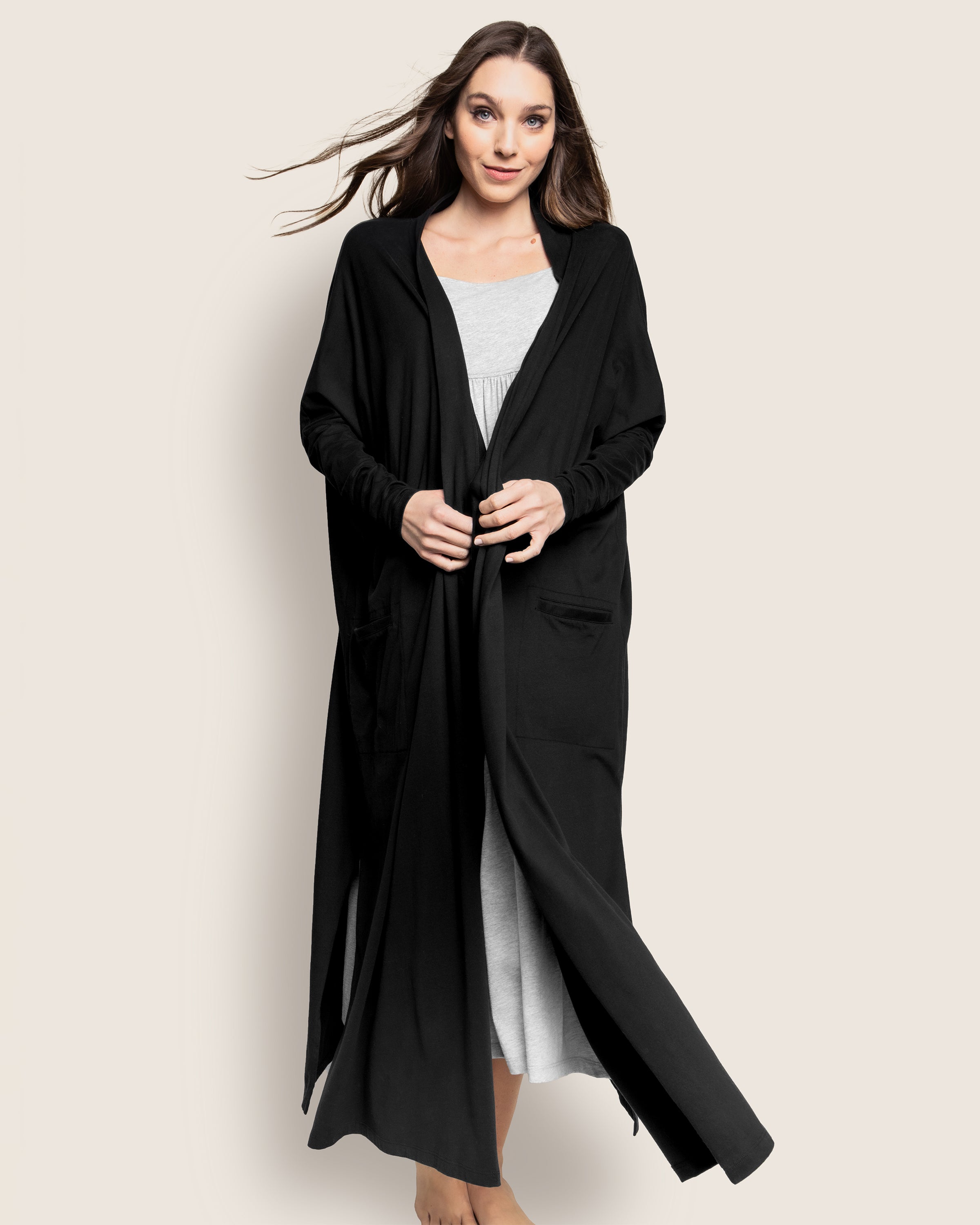 A woman models the Womens Pima Duster in Black by Petite Plume over a light gray jersey knit dress, her loose hair falling gently as she smiles slightly against a plain, light-toned background.