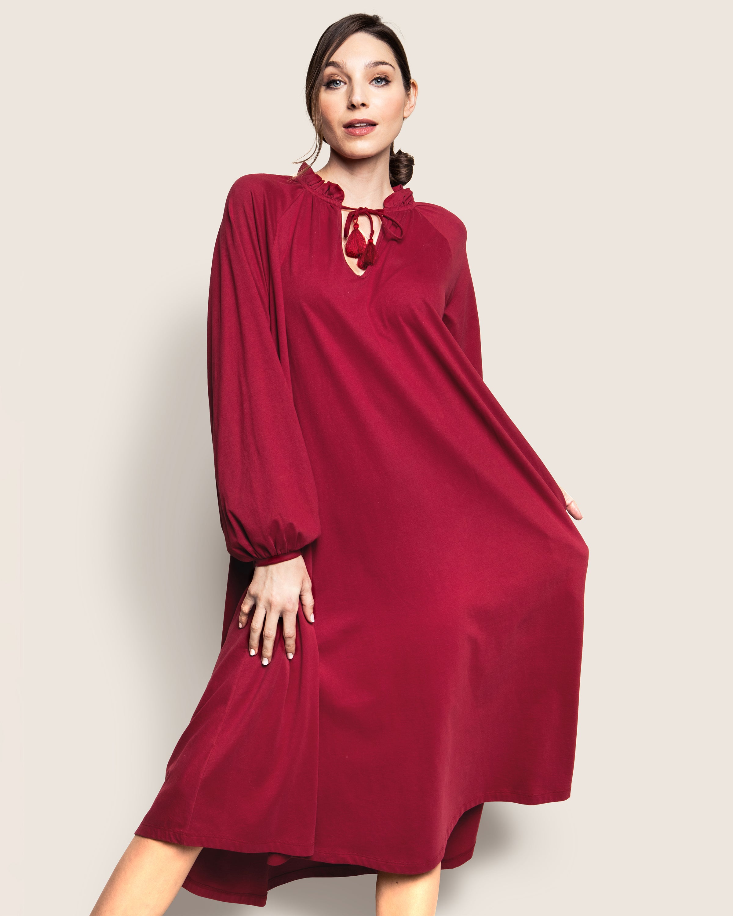 Against a plain backdrop, a person in Petite Plumes Womens Pima Garbo Nightgown in Bordeaux stands gracefully. The long-sleeved gown, featuring a bow-tied keyhole neckline, sways gently as they hold its sides with both hands.