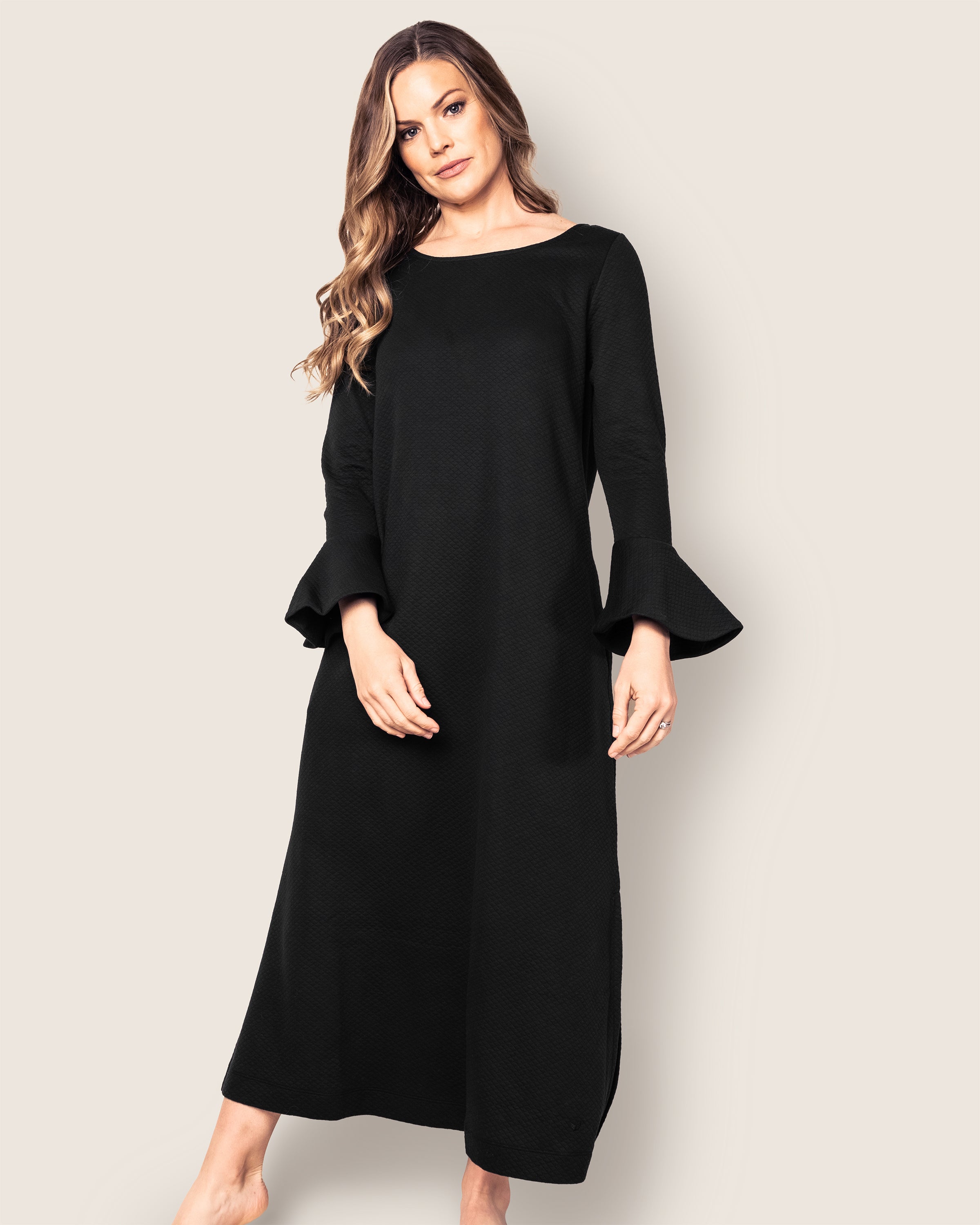 A person wearing the Womens Pima Ophelia Nightgown in Black by Petite Plume stands against a plain background. The luxurious gown, crafted from soft Pima cotton, elegantly drapes their form as their long, wavy hair cascades down, and they gaze directly at the camera.