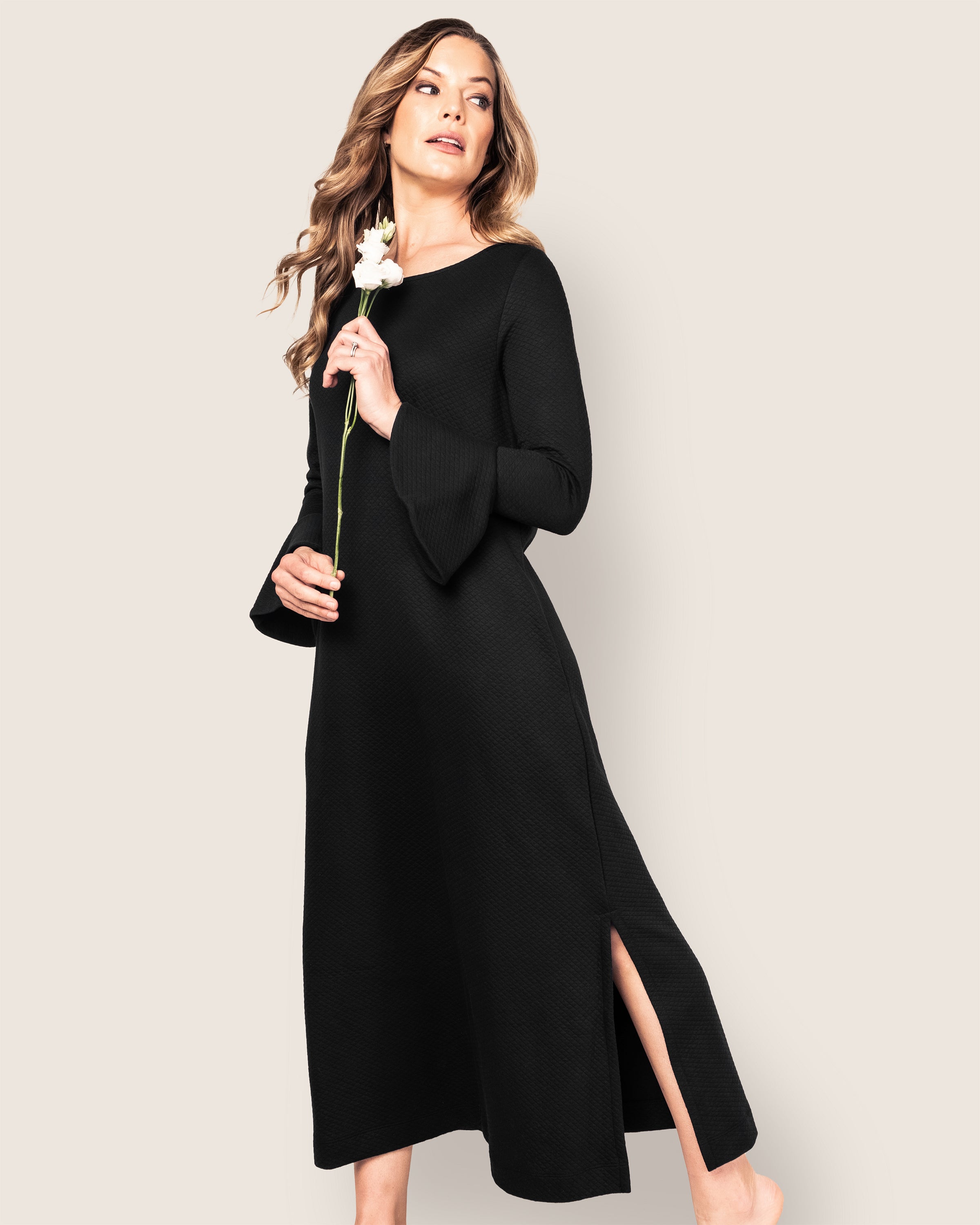 A woman in the Petite Plume Womens Pima Ophelia Nightgown in Black, featuring long sleeves and a side slit, holds a white flower against a neutral backdrop. She gazes upwards with a calm expression.