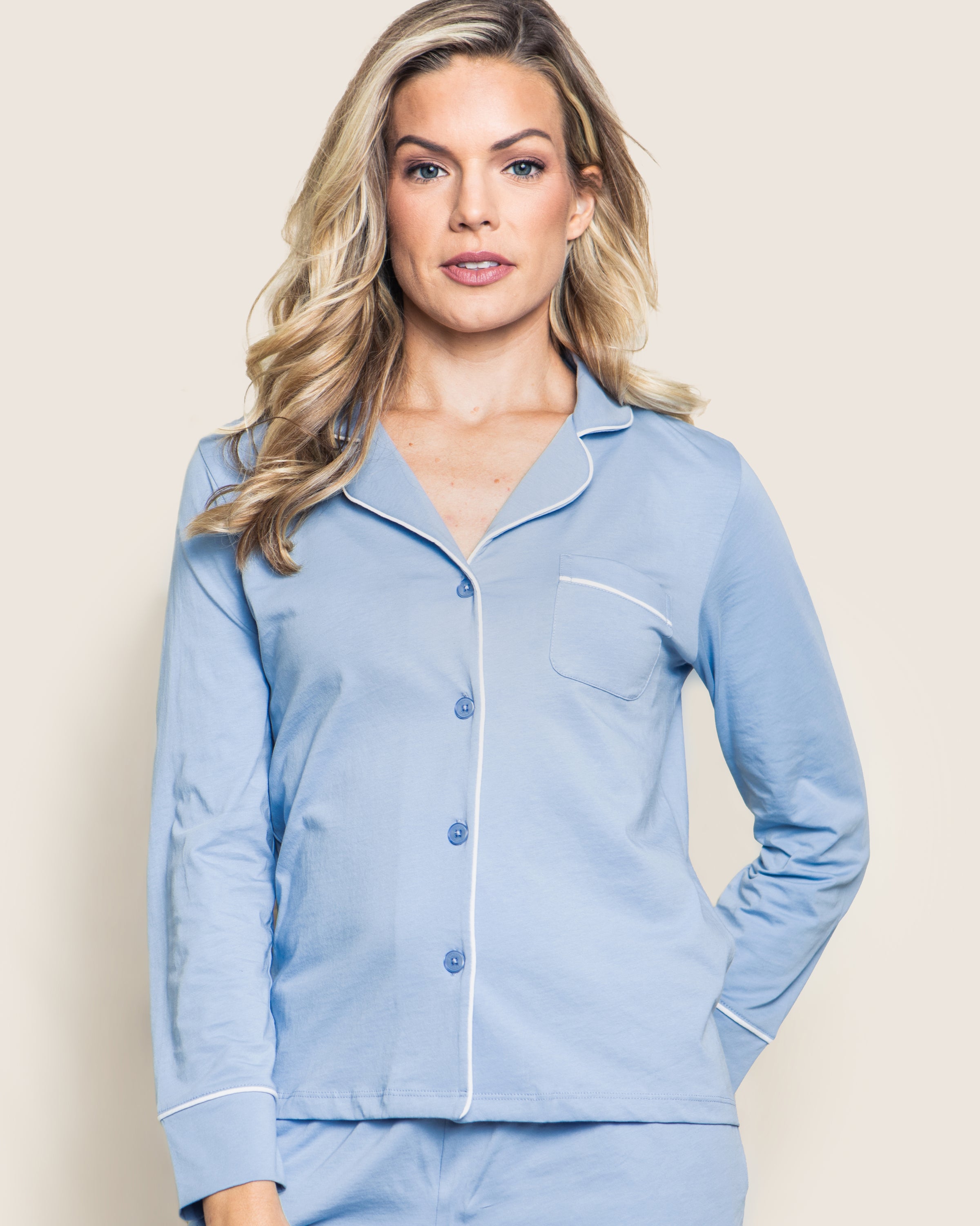 A person in Petite Plumes Womens Pima Pajama Short Set in periwinkle stands against a plain background. The luxury sleepwear features white piping, a single chest pocket, and blue buttons, complementing their long, wavy blonde hair.