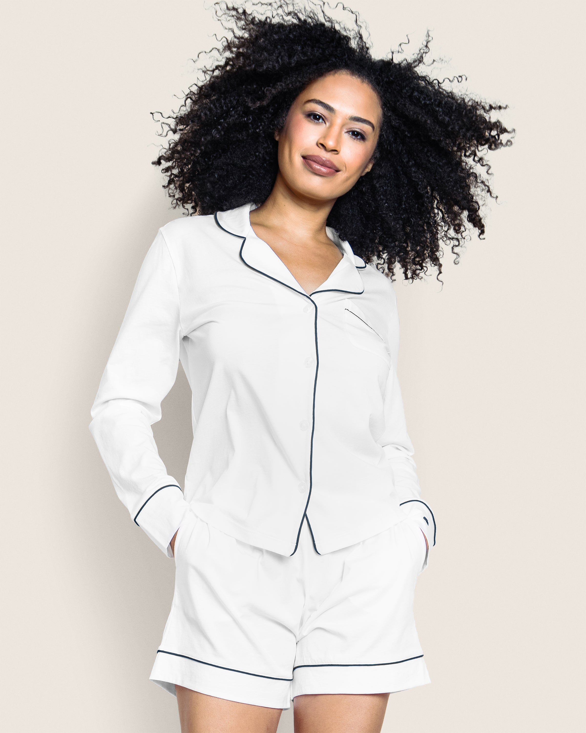 A person with curly hair smiles in Petite Plumes Womens Pima Long Sleeve Short Set, made from Peruvian Pima cotton. The luxurious white pajamas with black piping and shorts pockets exude comfort as they pose against a light beige background.