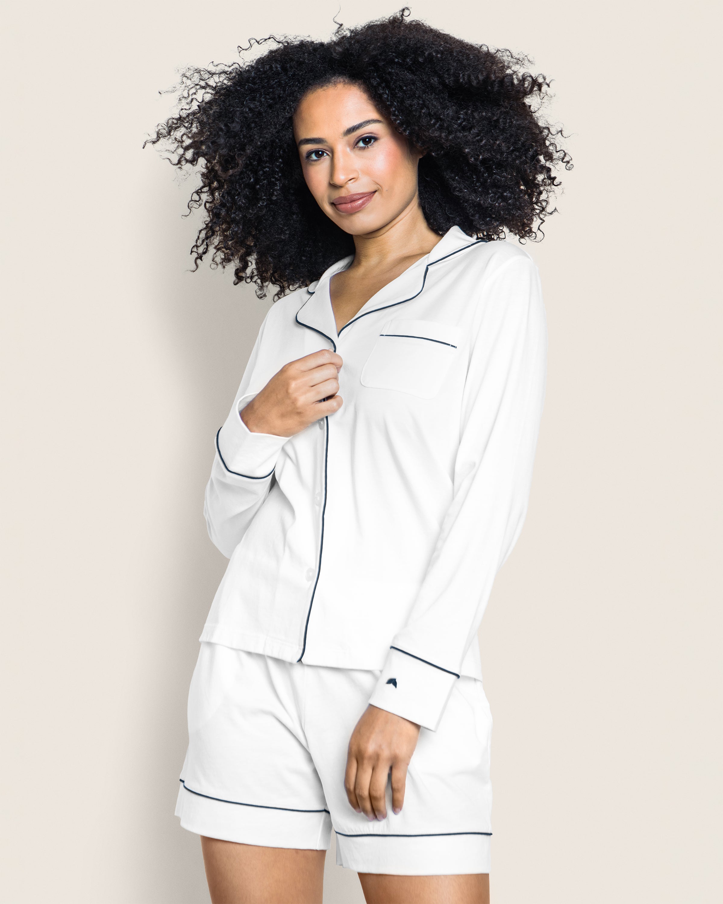A person with curly hair poses against a plain background wearing the luxurious Petite Plume Womens Pima Long Sleeve Short Set in white with black piping, gently touching the collar with a smile.